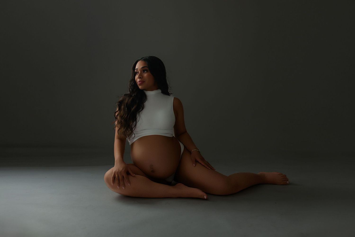 Pregnant woman in white bodysuit sitting gracefully on studio floor, showcasing baby bump in elegant maternity photography session.