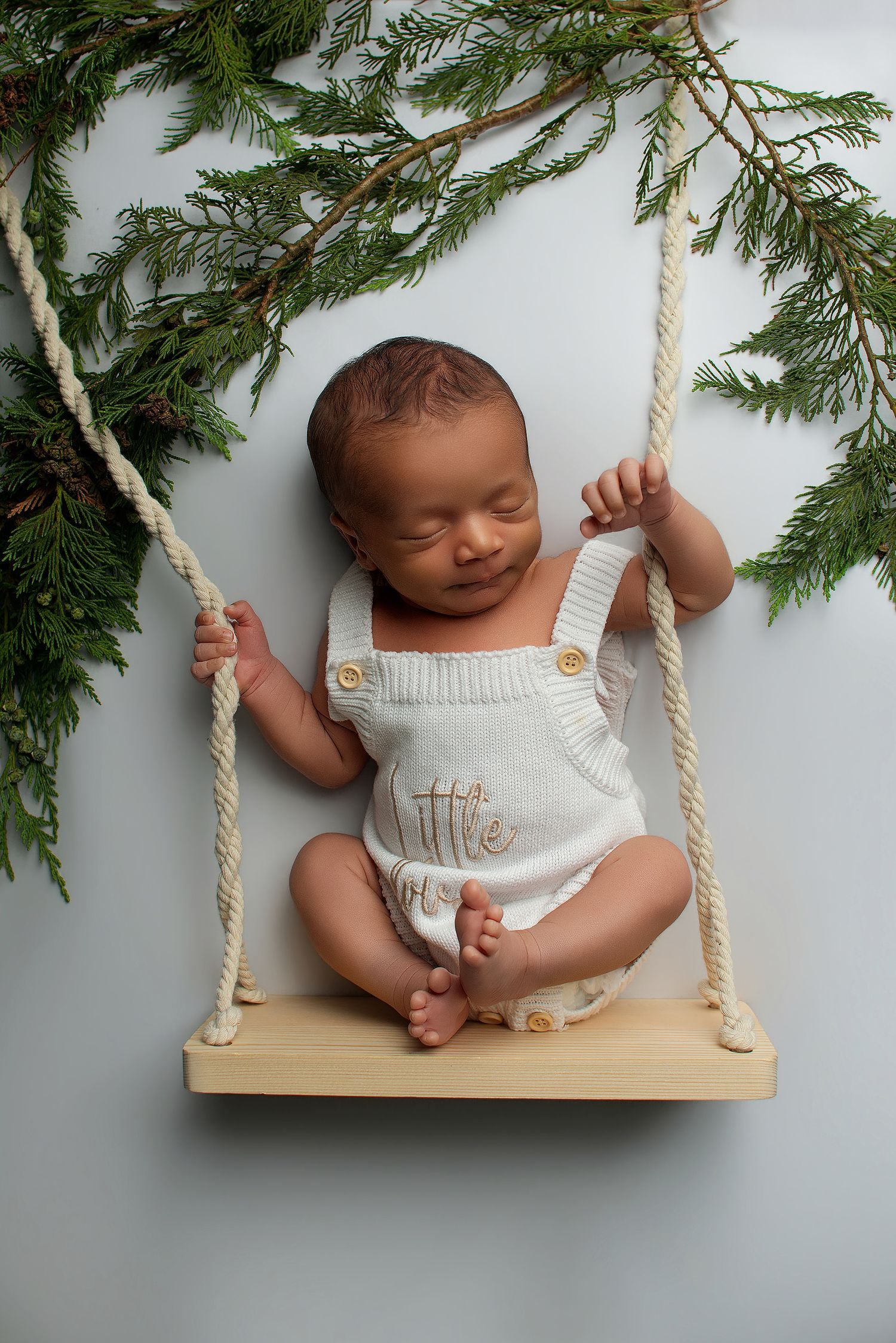 Sleeping newborn baby in white knit overalls posed on a wooden rope swing with green pine branches.