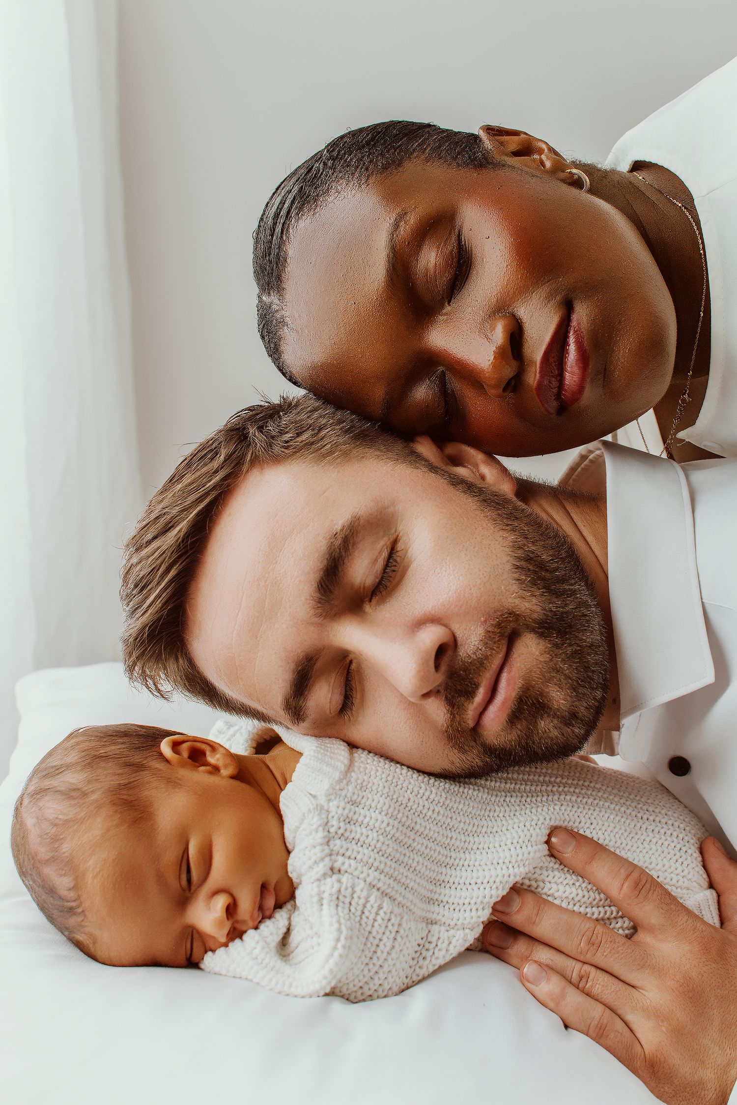 Interracial family with sleeping newborn baby, parents resting with eyes closed on white bedding.