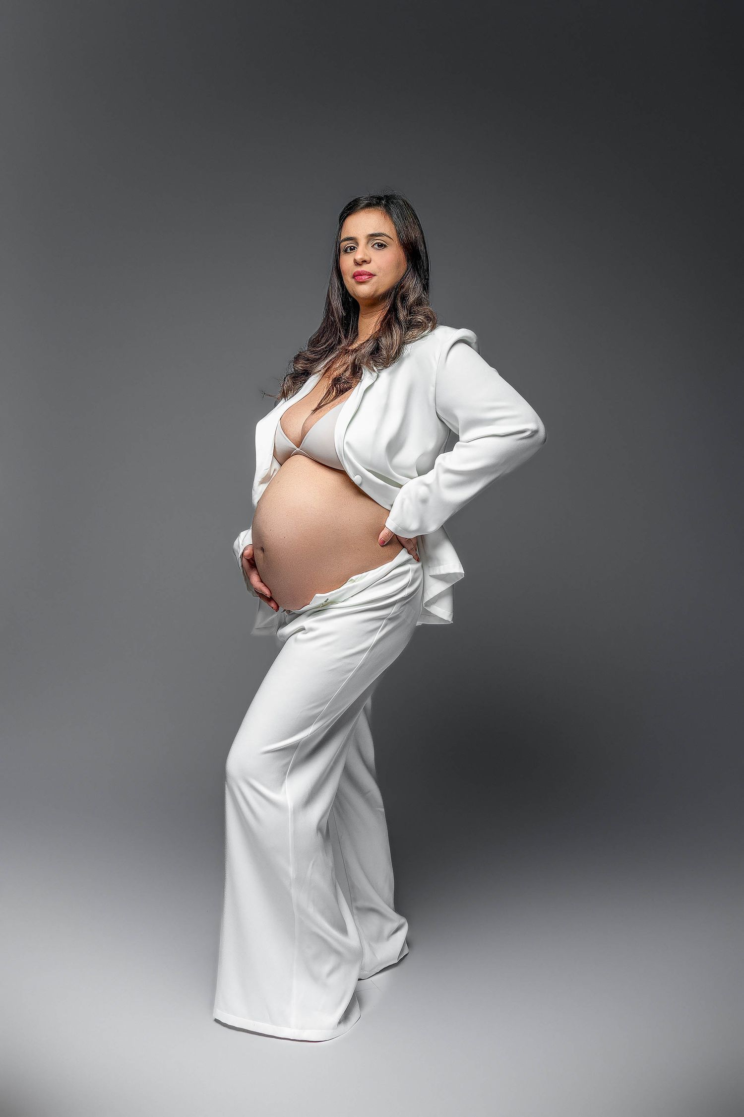 Elegant pregnant woman in white blazer and flared pants cradling her baby bump on gray background.