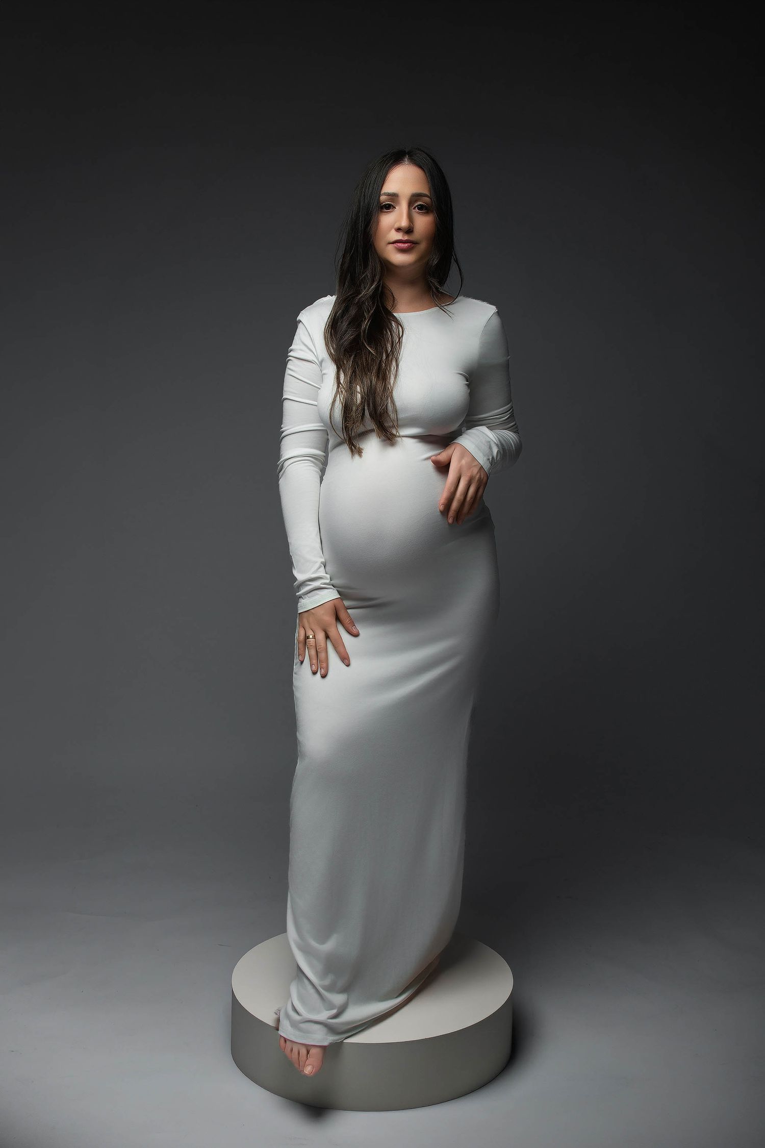 Pregnant woman in white long-sleeve fitted maternity gown standing on circular platform against gray studio background.