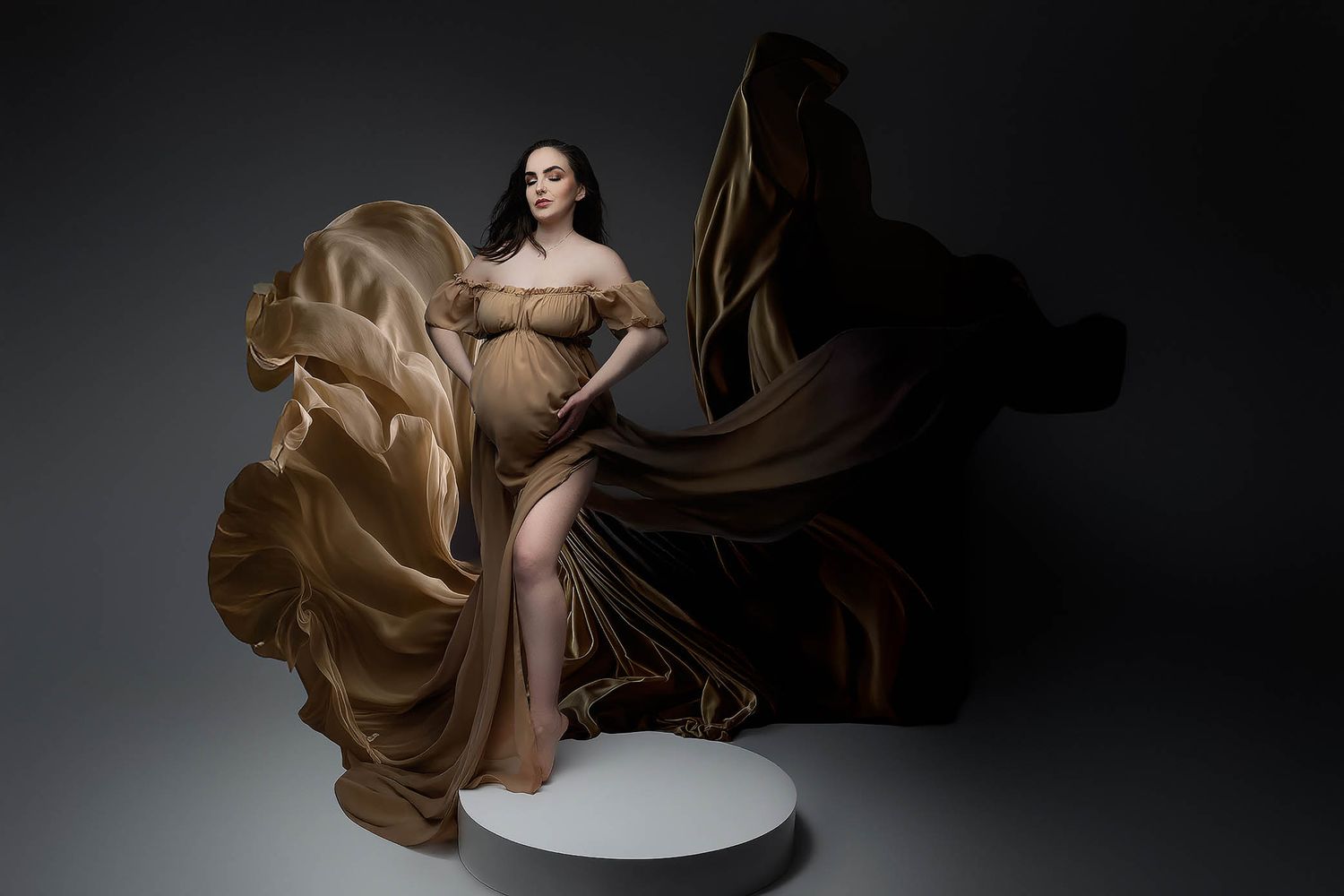 Pregnant woman in flowing golden gown with dramatic fabric movement against dark studio background.