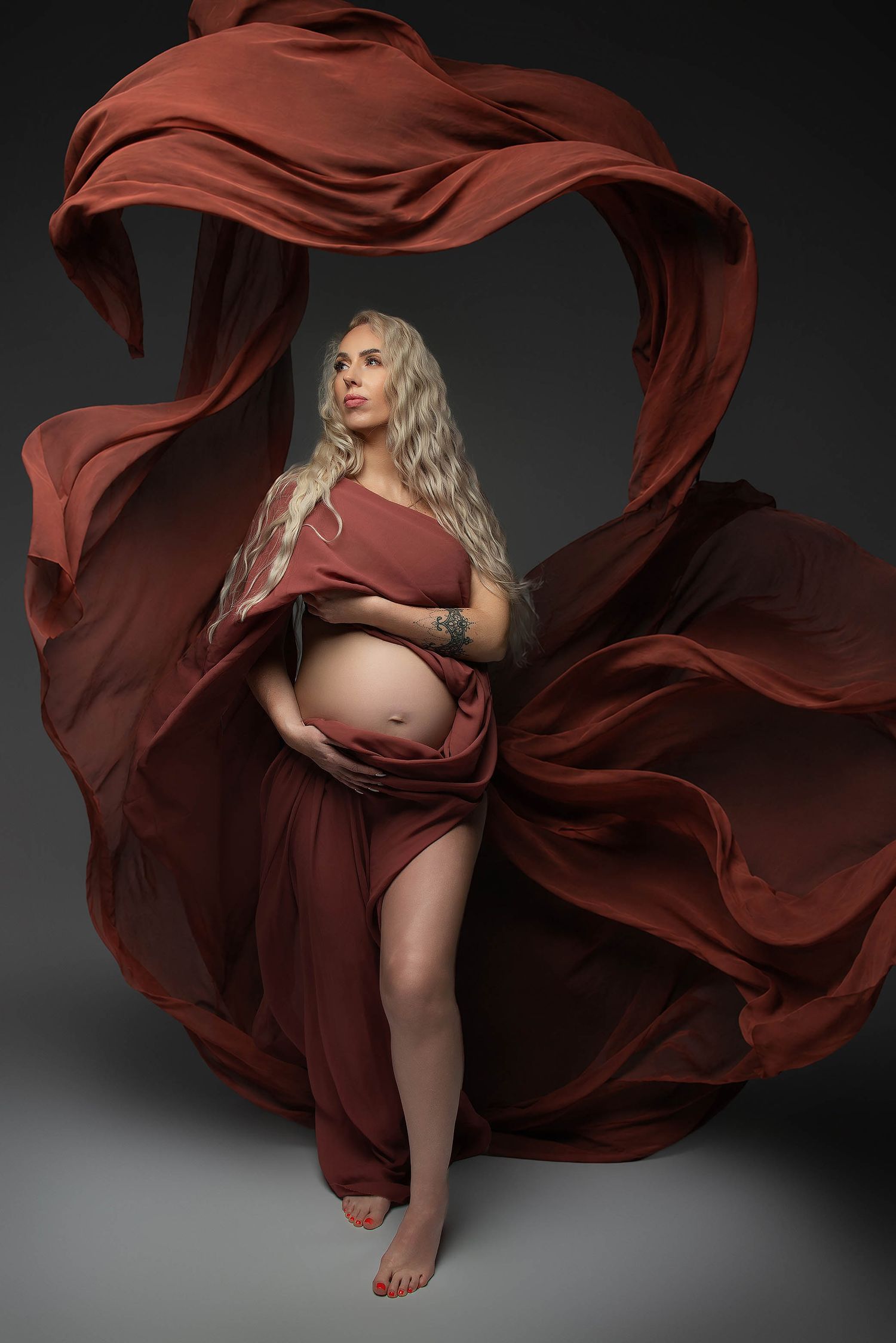 Pregnant woman draped in flowing rust-red fabric, showcasing her baby bump in an elegant maternity studio portrait.