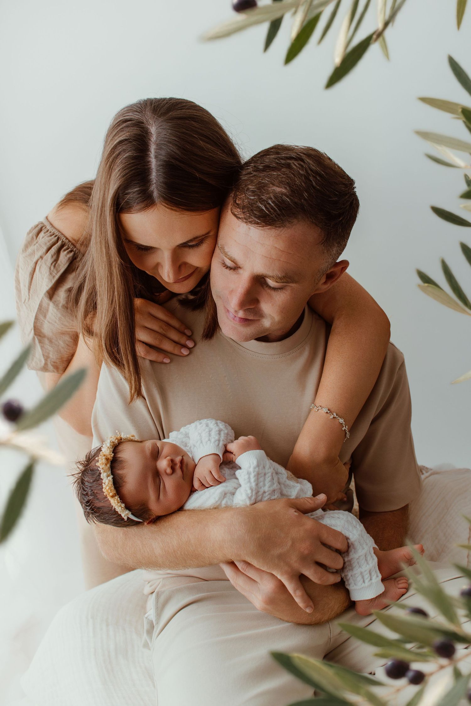 Young couple tenderly holding their sleeping newborn baby, surrounded by olive branches in a soft studio setting.