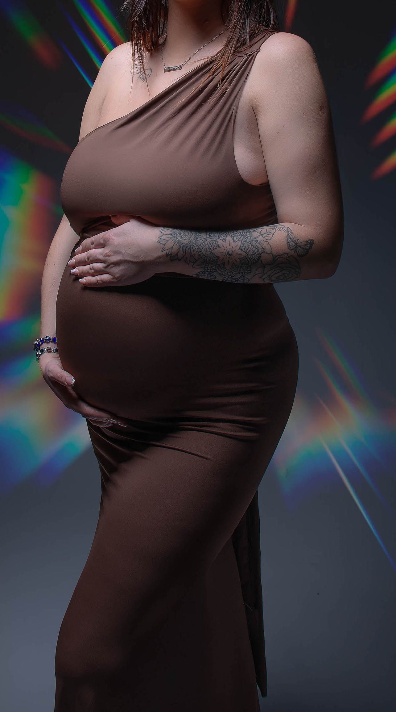 Pregnant woman in brown one-shoulder maxi dress cradling baby bump, with colorful light refractions in background.