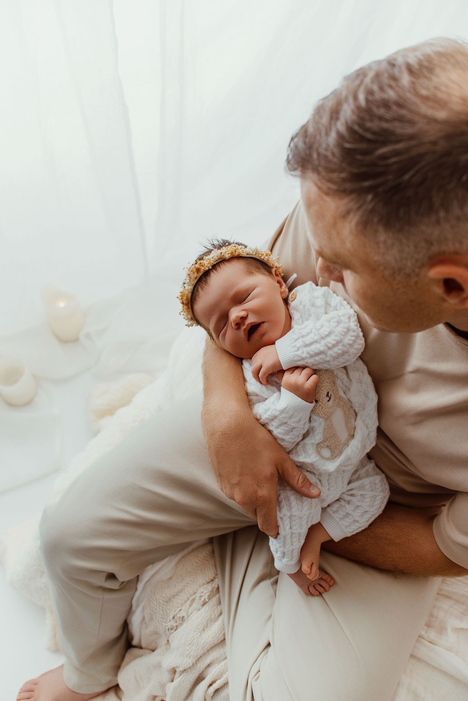 A father tenderly holds his newborn baby girl wearing a gold headband in a soft, white-lit setting.