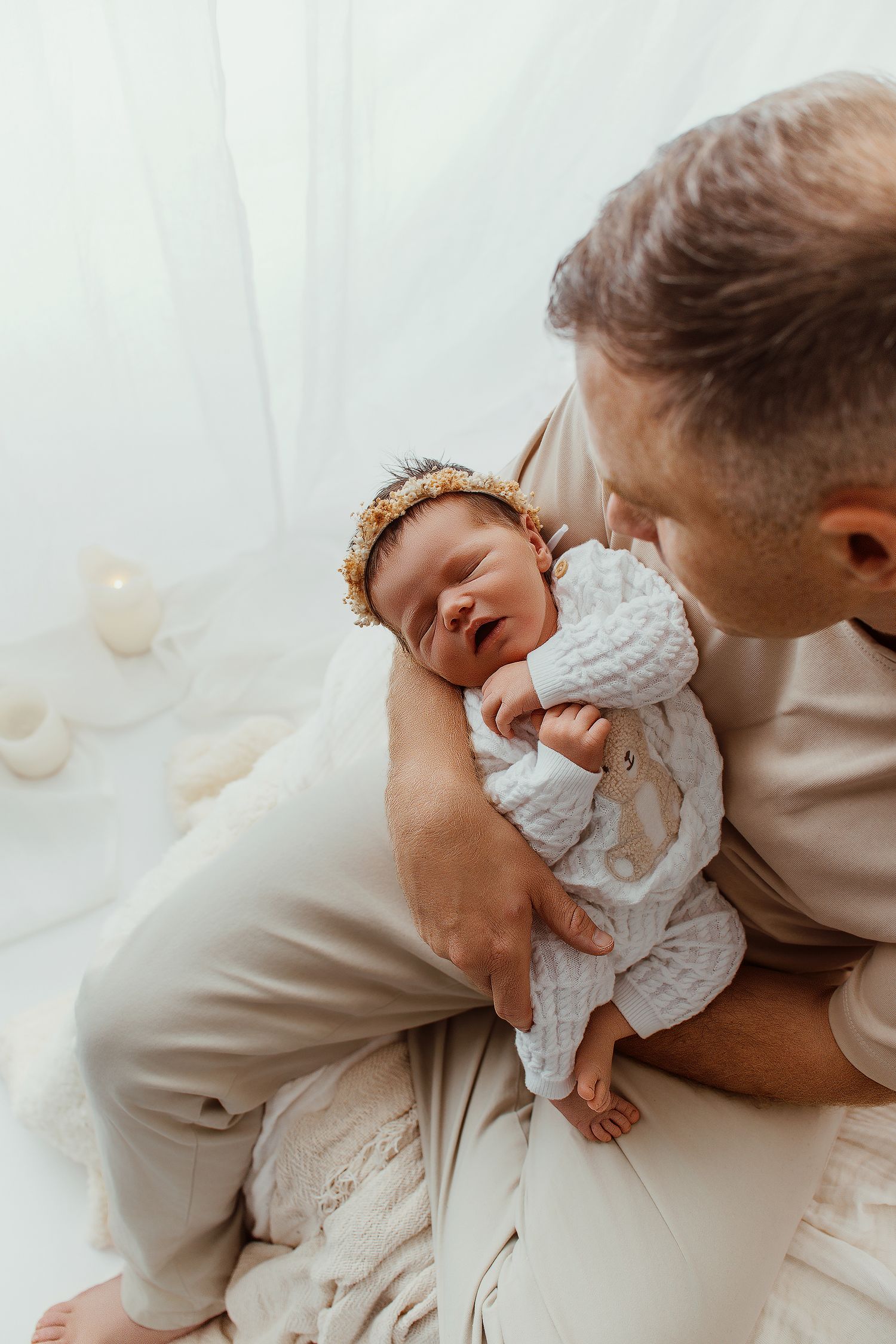 Father cradling newborn baby girl wearing gold floral headband in white studio setting.