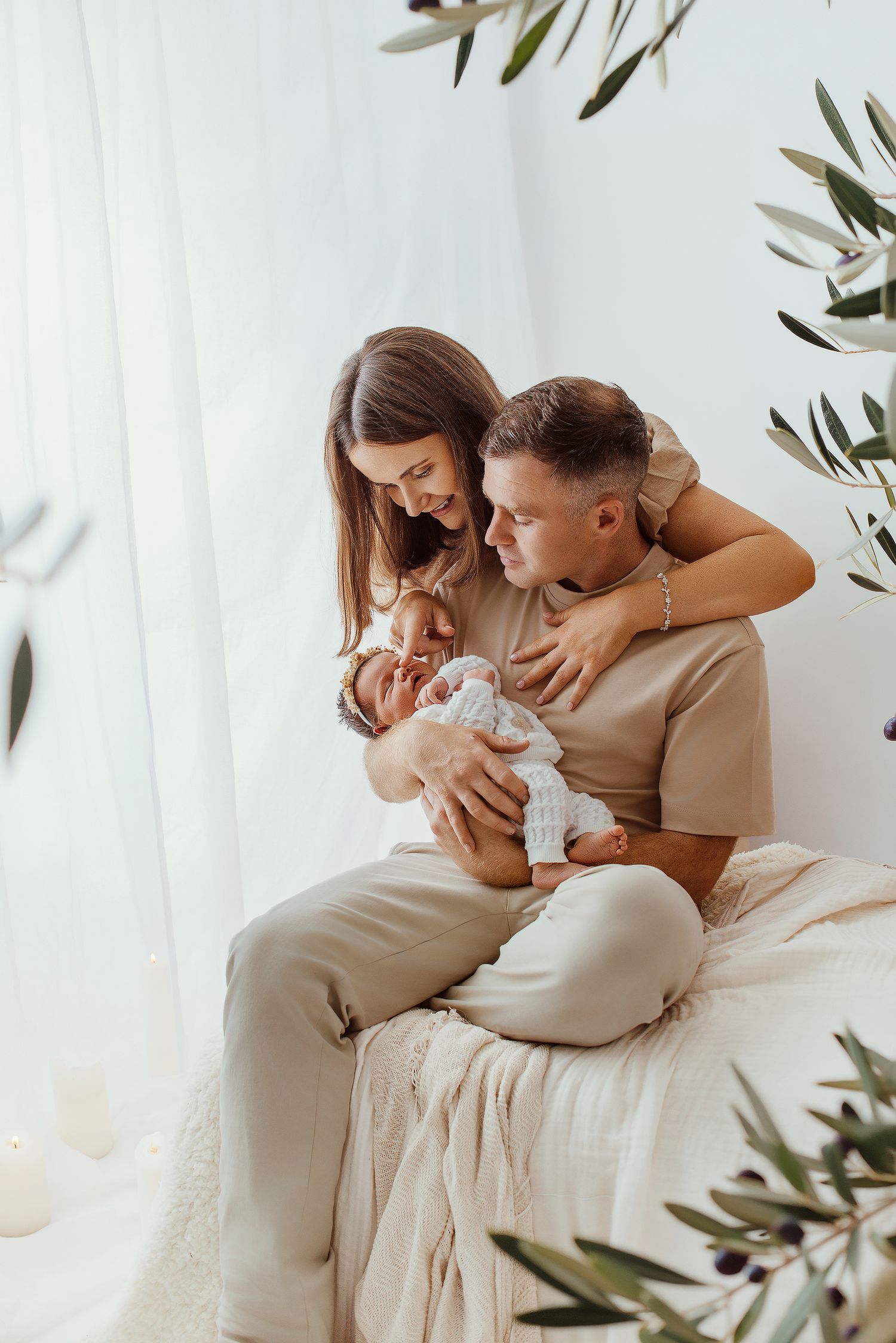 A young couple in beige outfits sitting together, lovingly cradling their newborn baby in a bright, serene room.