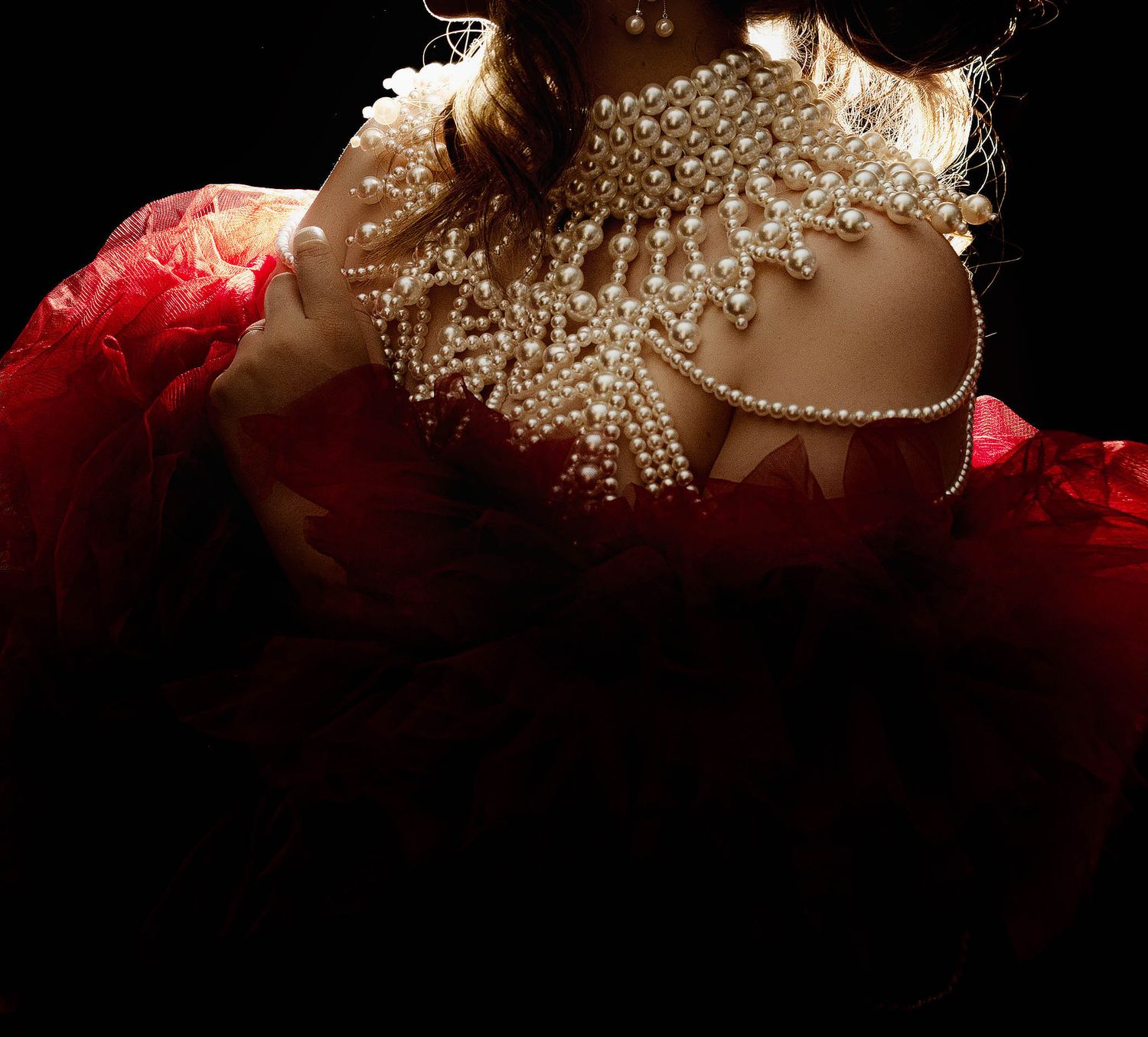 Woman in red dress with elaborate pearl embellishment across shoulders and neckline against dark background.