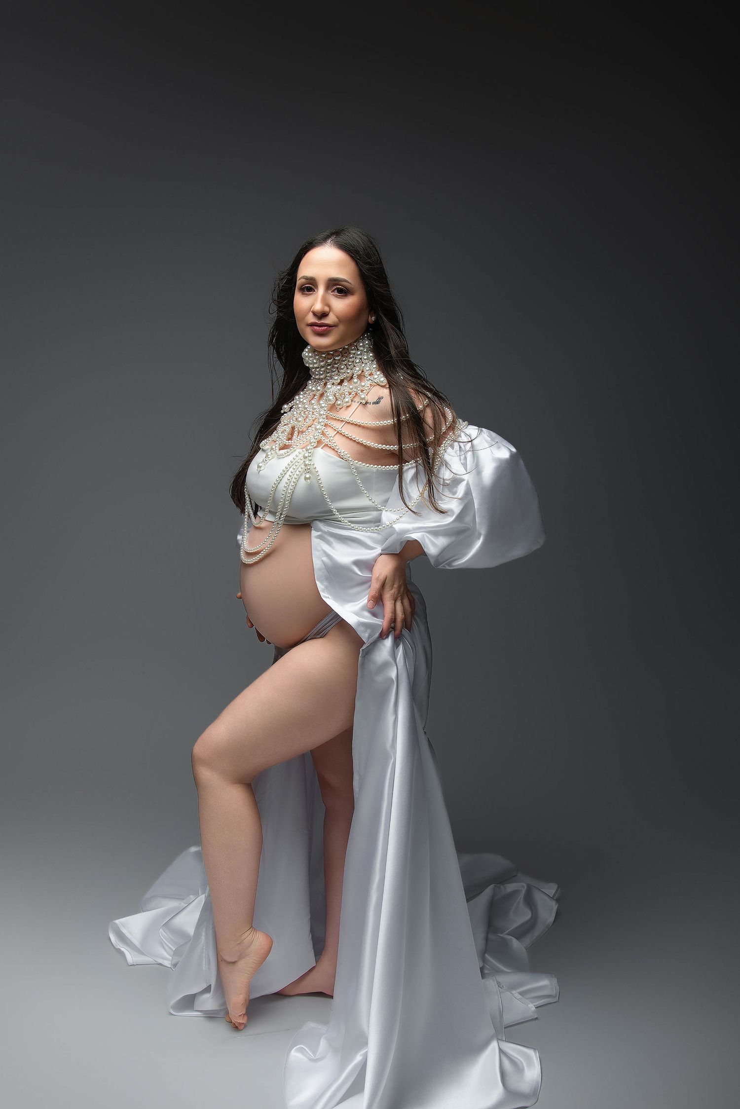 Pregnant woman in white satin robe and pearl jewelry poses gracefully in a professional maternity photo shoot.
