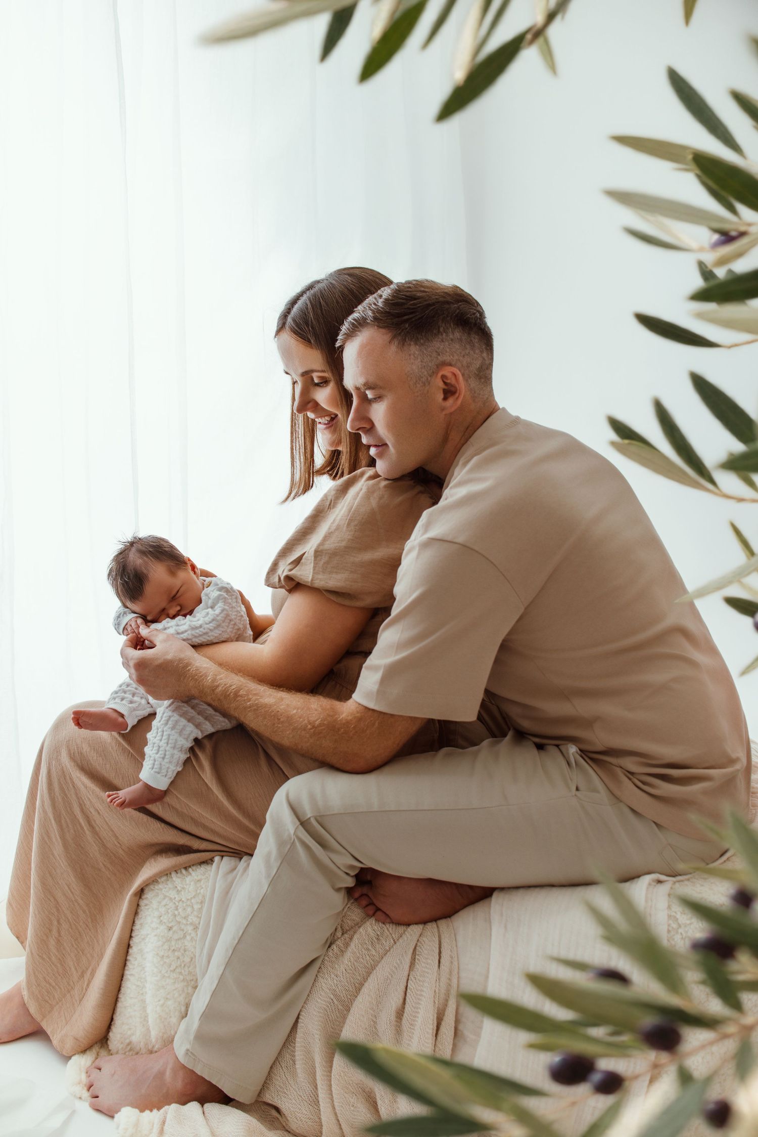 A couple sits together on a bed, lovingly gazing at their newborn baby in a bright, minimalist room.