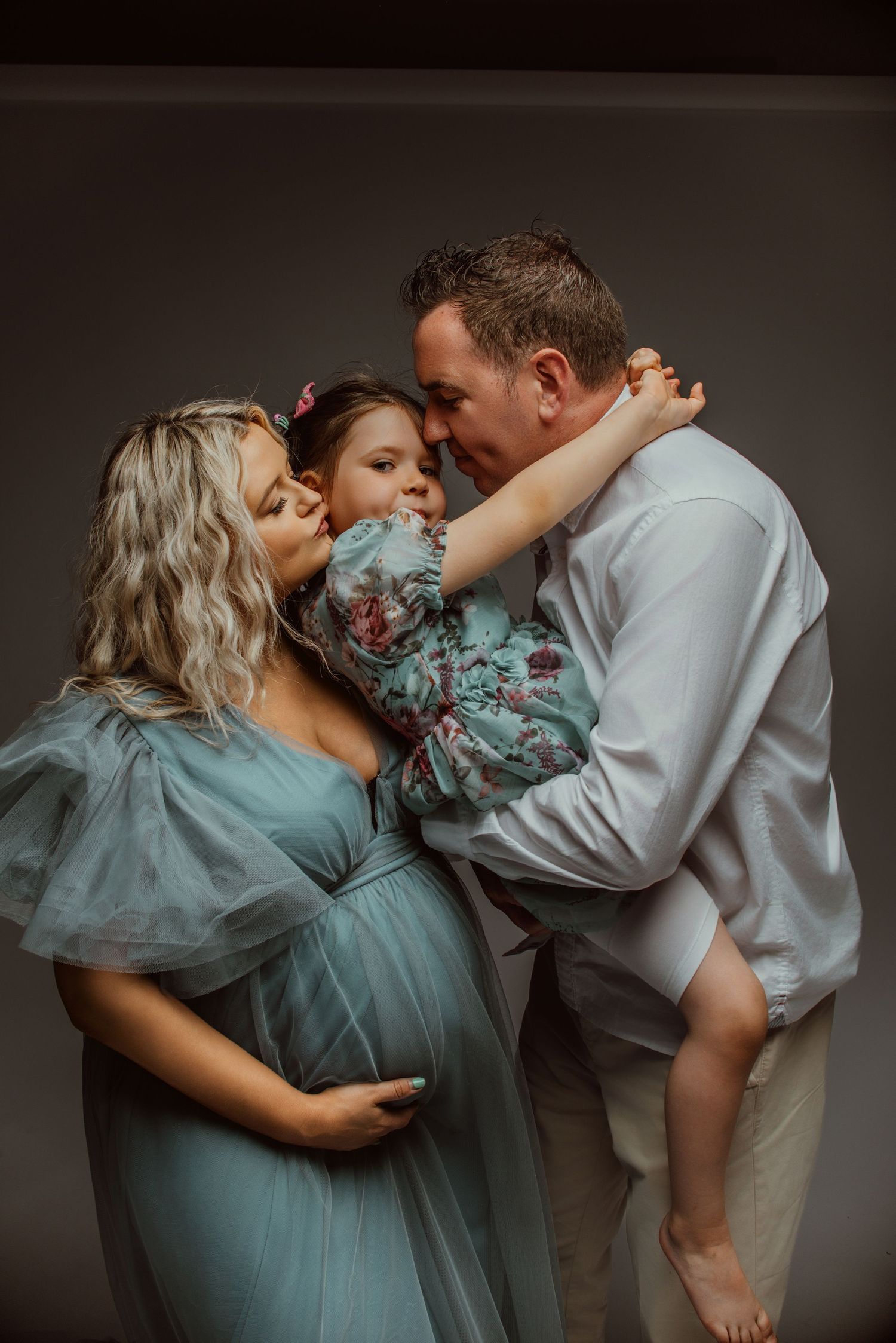 Pregnant woman in teal gown with husband holding daughter during maternity photo session.