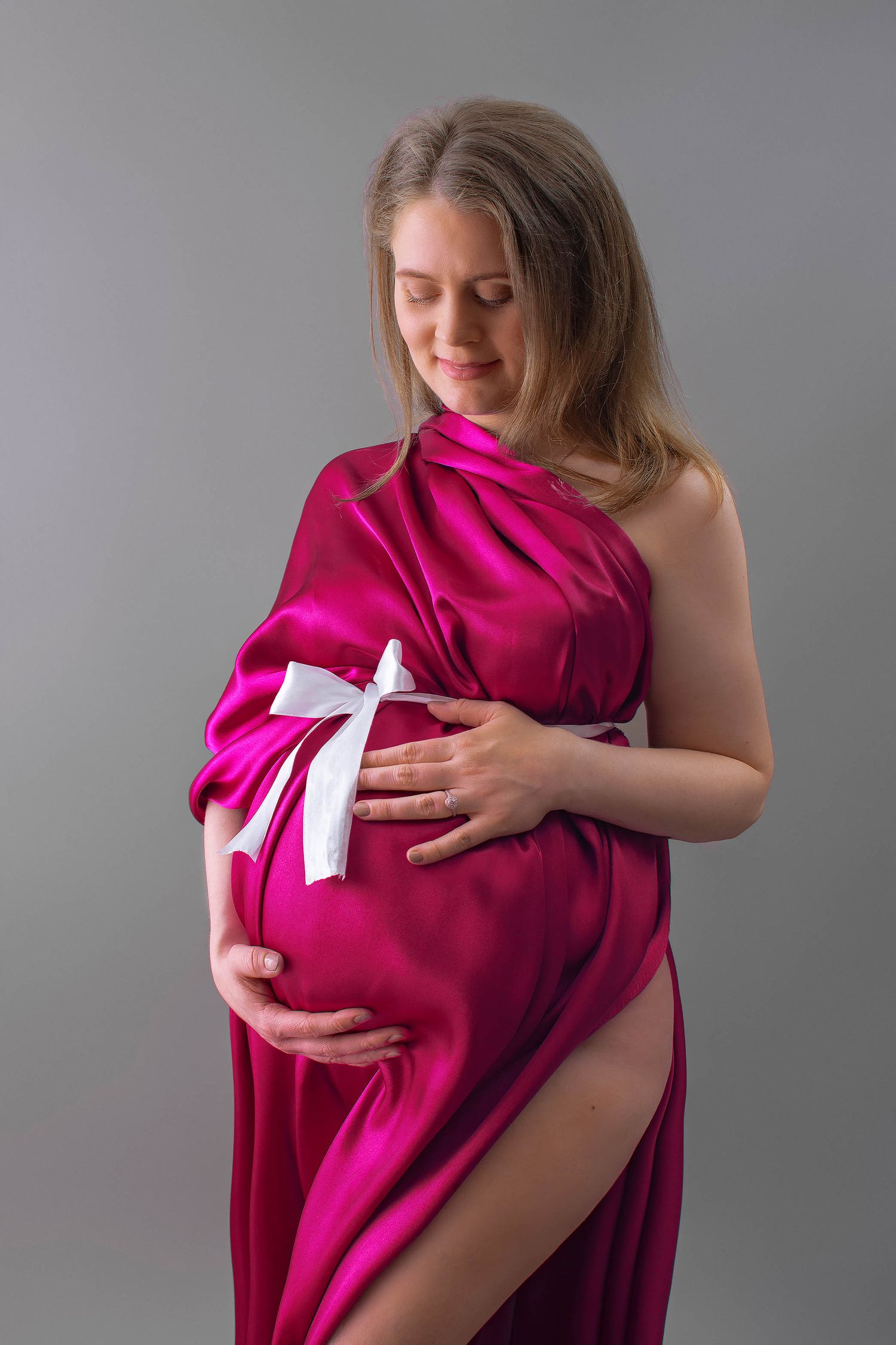 Pregnant woman in pink satin fabric with white bow, cradling baby bump in studio.