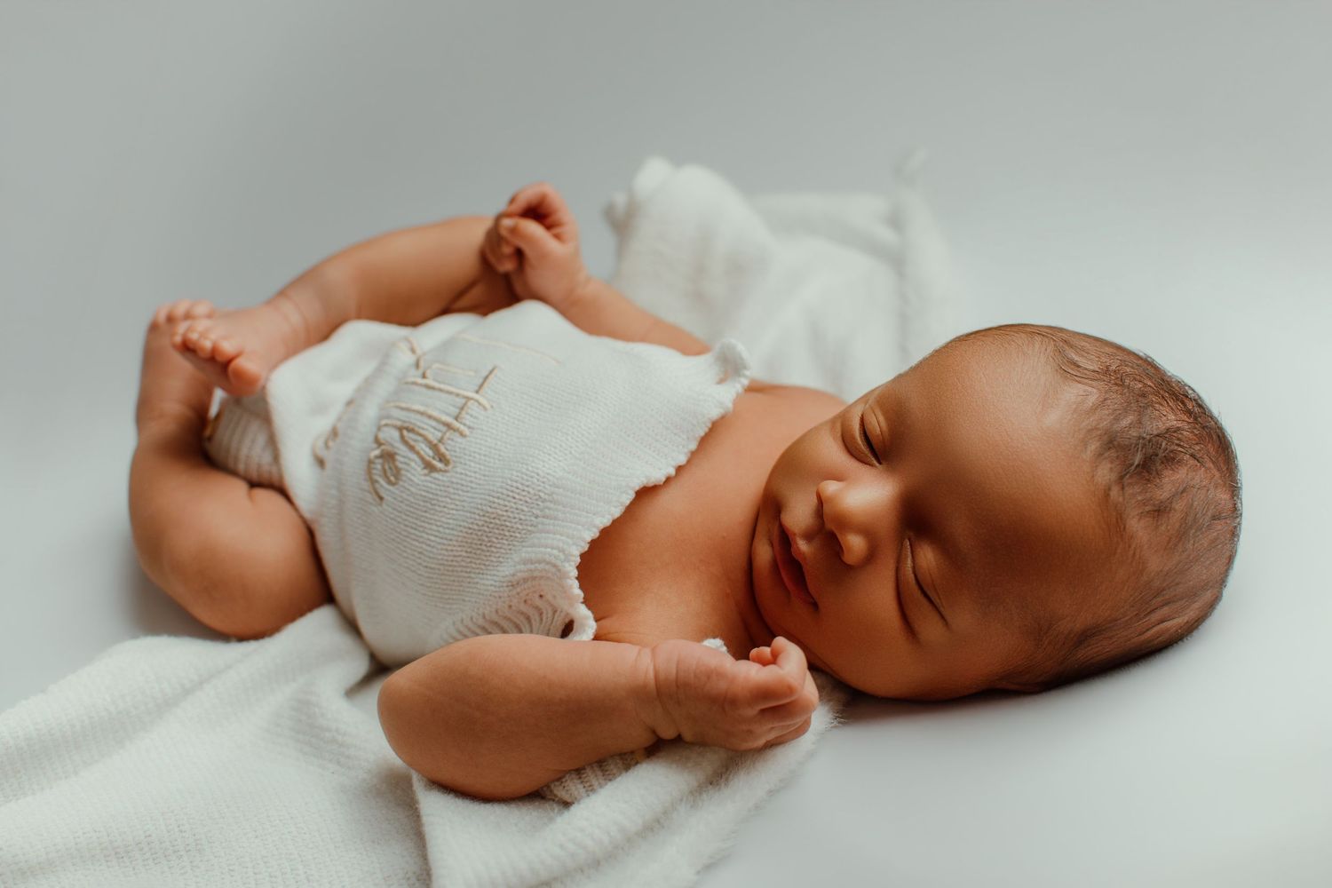 Peaceful newborn baby sleeping on a white blanket wearing a white onesie, with tiny hands and feet visible.