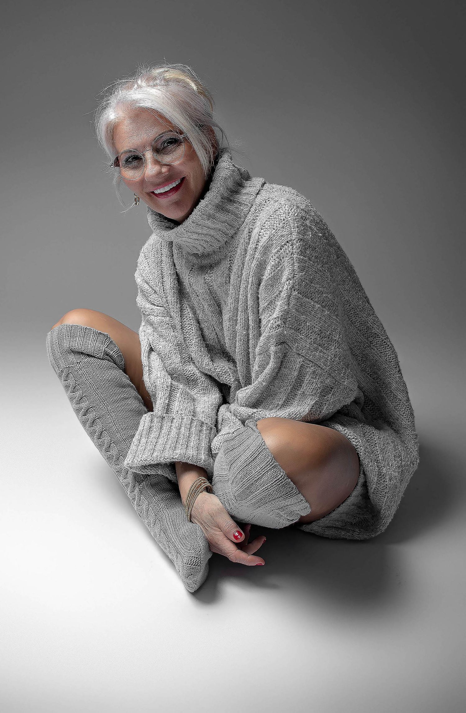 Smiling older woman with gray hair sitting cross-legged in cozy gray oversized turtleneck sweater and knee-high socks.