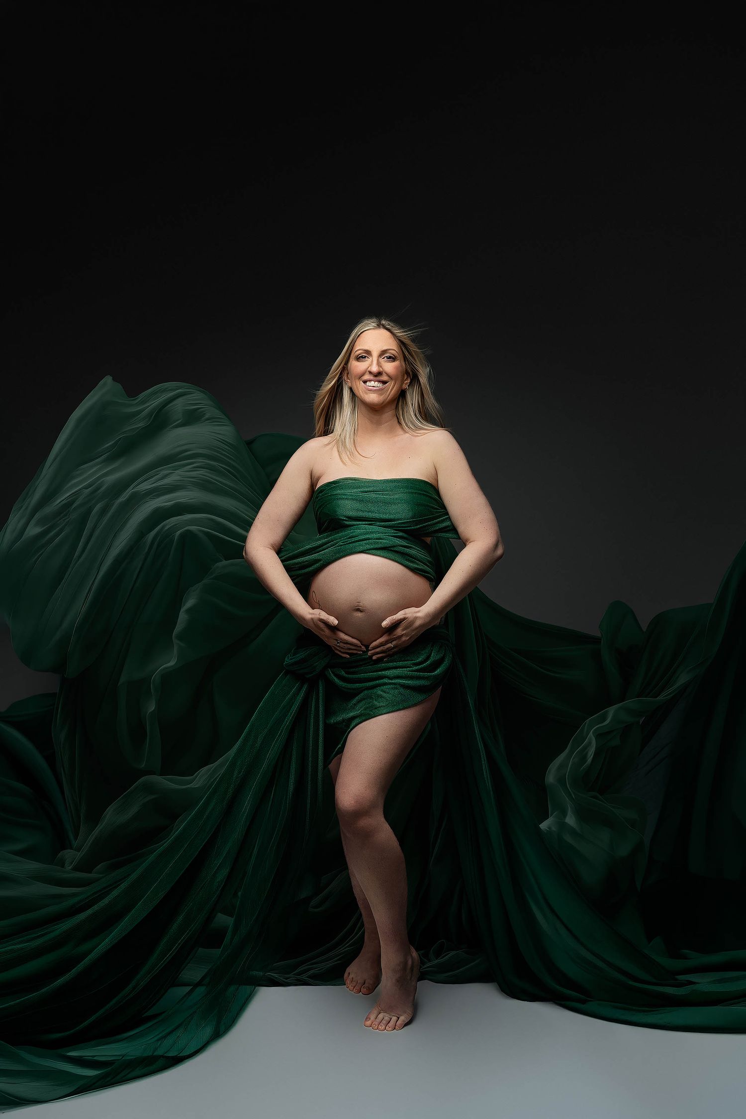 Smiling pregnant woman in flowing emerald green fabric wrap poses barefoot against dark studio background.