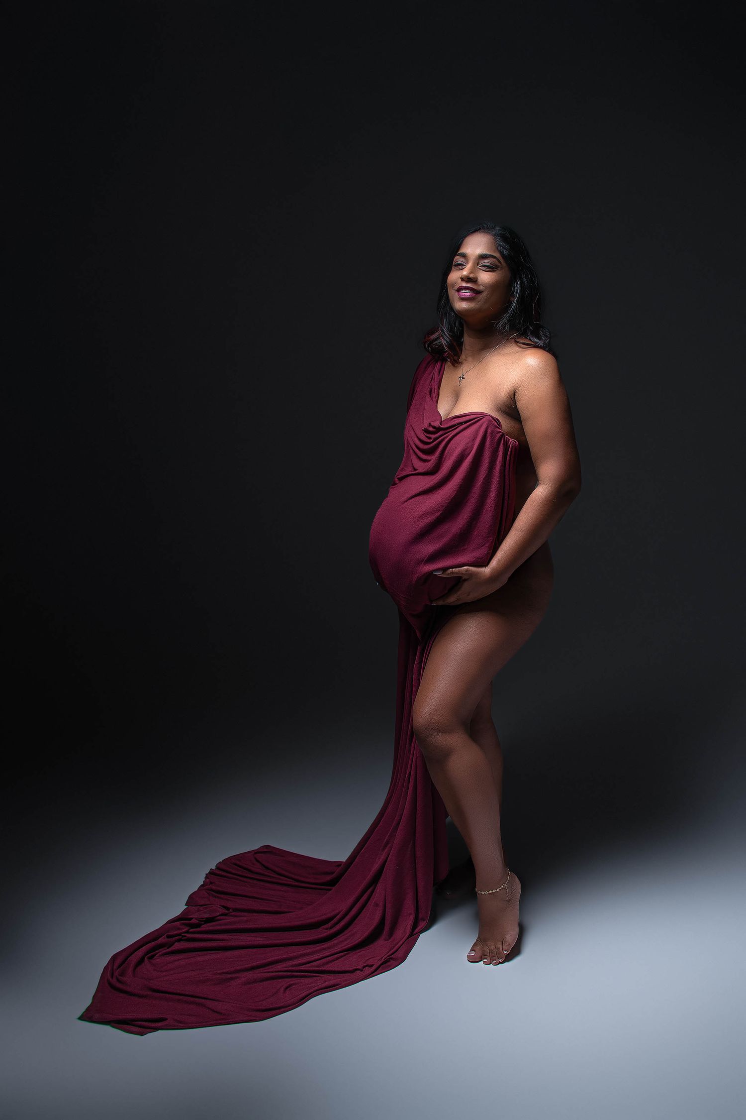 Pregnant woman draped in elegant burgundy fabric poses gracefully against dark studio background.