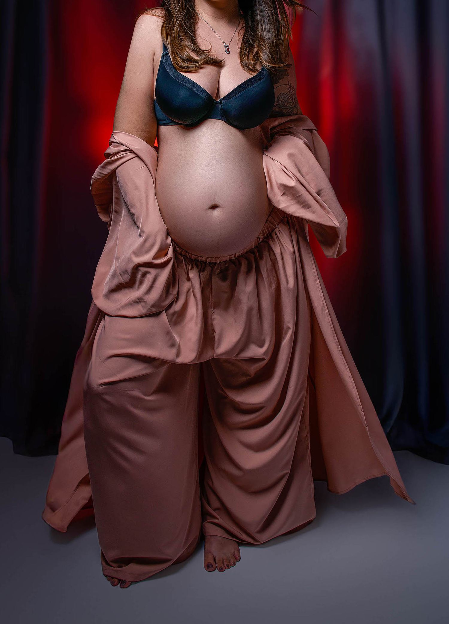 Pregnant woman in teal bra and rose satin outfit cradling bare baby bump against dramatic red backdrop.