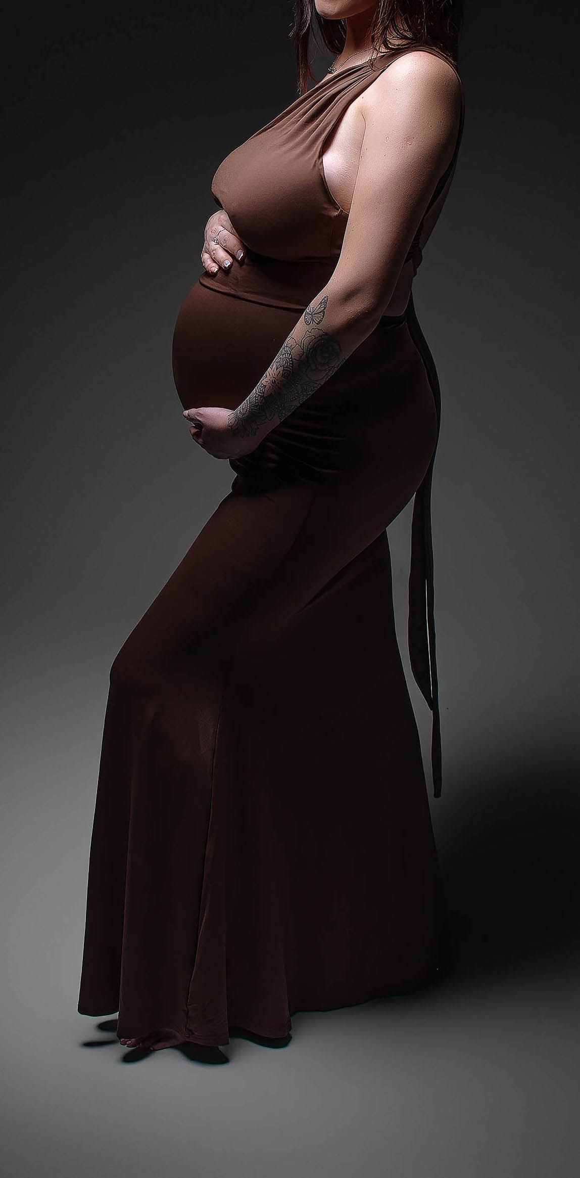 Pregnant woman in elegant brown halter maxi dress cradling baby bump, showcasing maternity fashion in studio portrait.