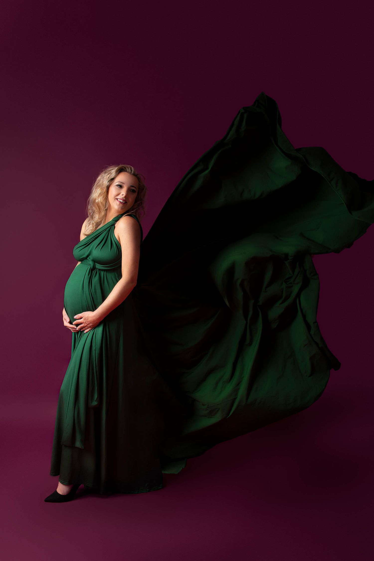 Pregnant woman in flowing emerald green gown with dramatic fabric wings poses against deep magenta studio background.
