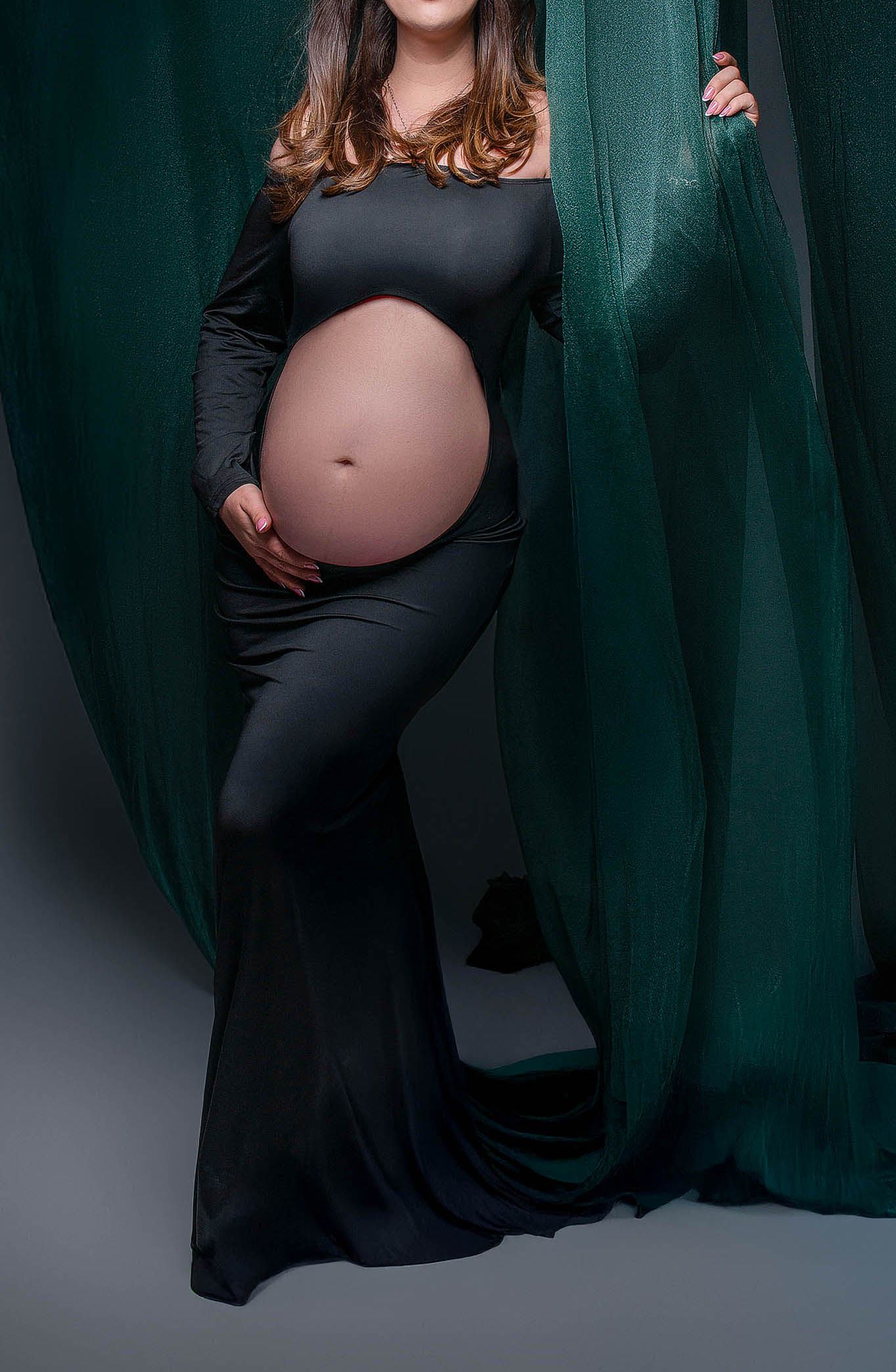 Pregnant woman in black off-shoulder gown holding baby bump, posed between emerald green drapes in maternity photo.