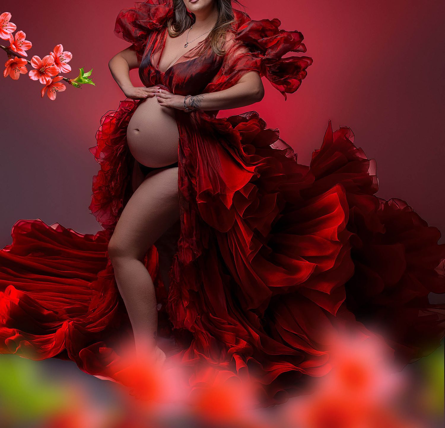 Pregnant woman in flowing red ruffled maternity gown poses gracefully among red flowers in a studio setting.