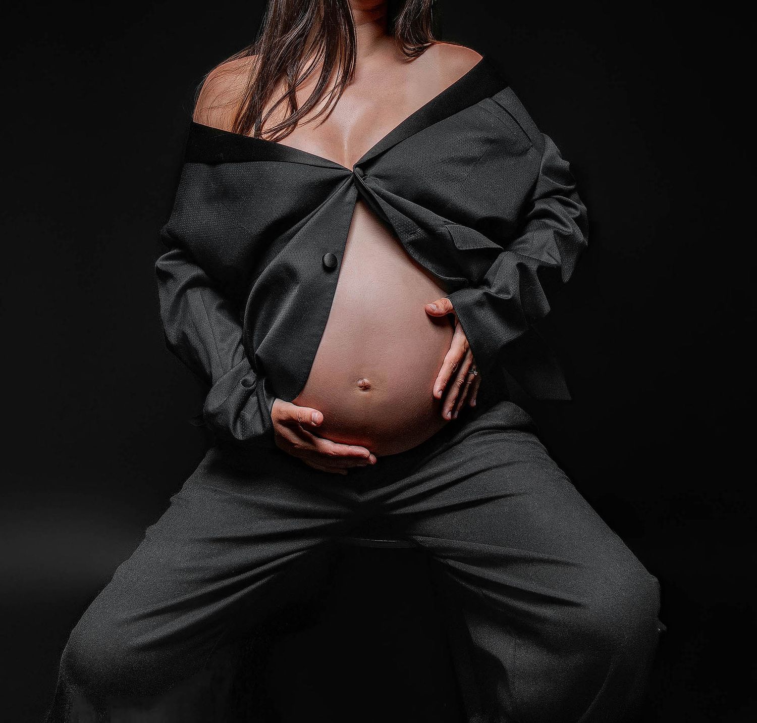 Pregnant woman in black off-shoulder top and pants cradling baby bump against dark background.
