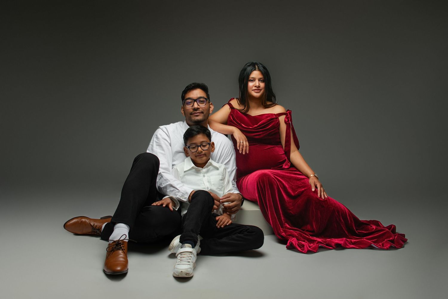 Pregnant woman in red velvet gown poses with husband and child in studio maternity family portrait.