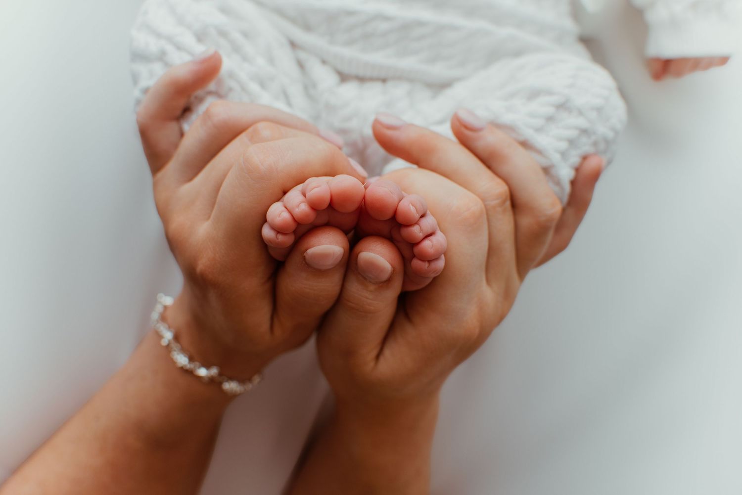 Close-up of a mother's hands gently cradling tiny newborn baby feet, wearing a delicate pearl bracelet.