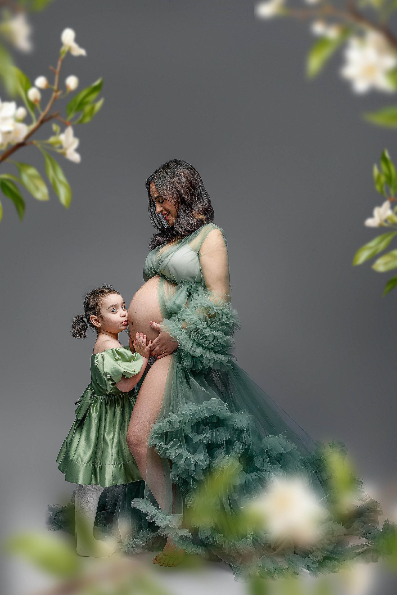 Pregnant woman in green tulle gown with young daughter hugging her belly amid white flowers.