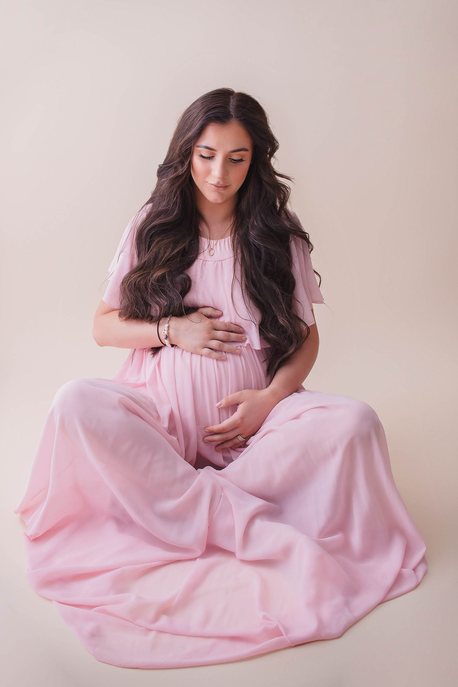 Pregnant woman in flowing pink dress sitting gracefully, cradling baby bump with long wavy dark hair.