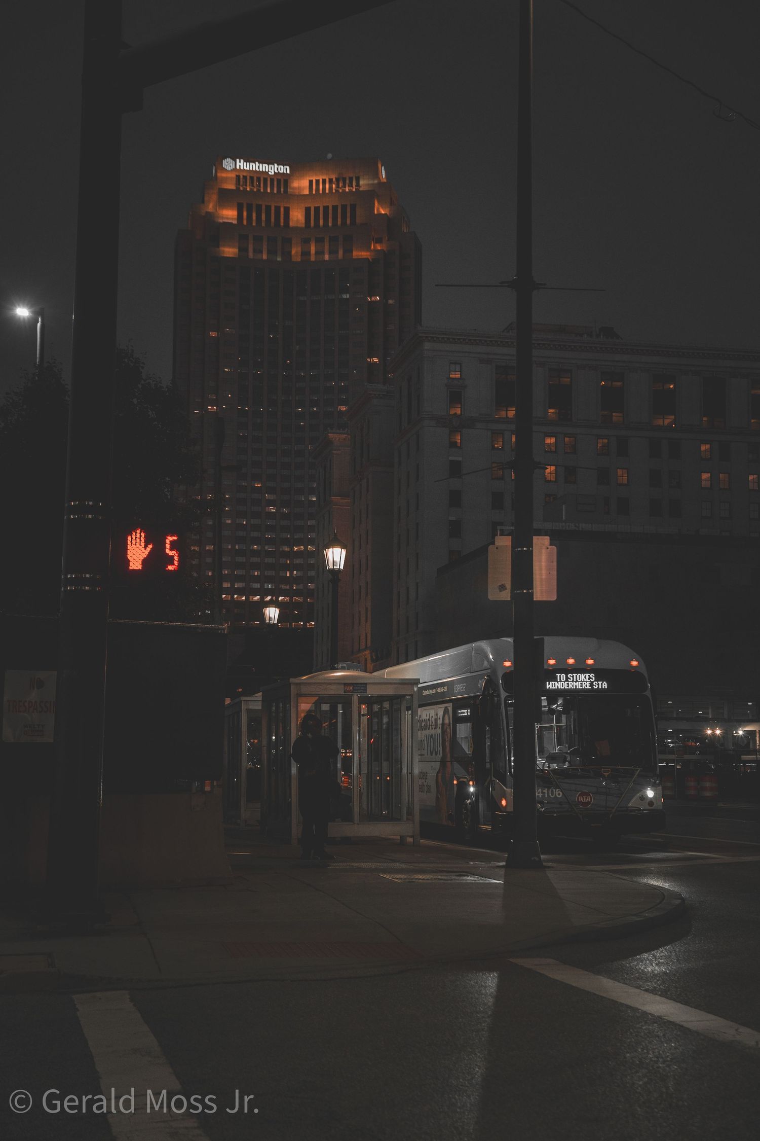 Nighttime urban street scene with illuminated building and bus stop in warm orange tones.