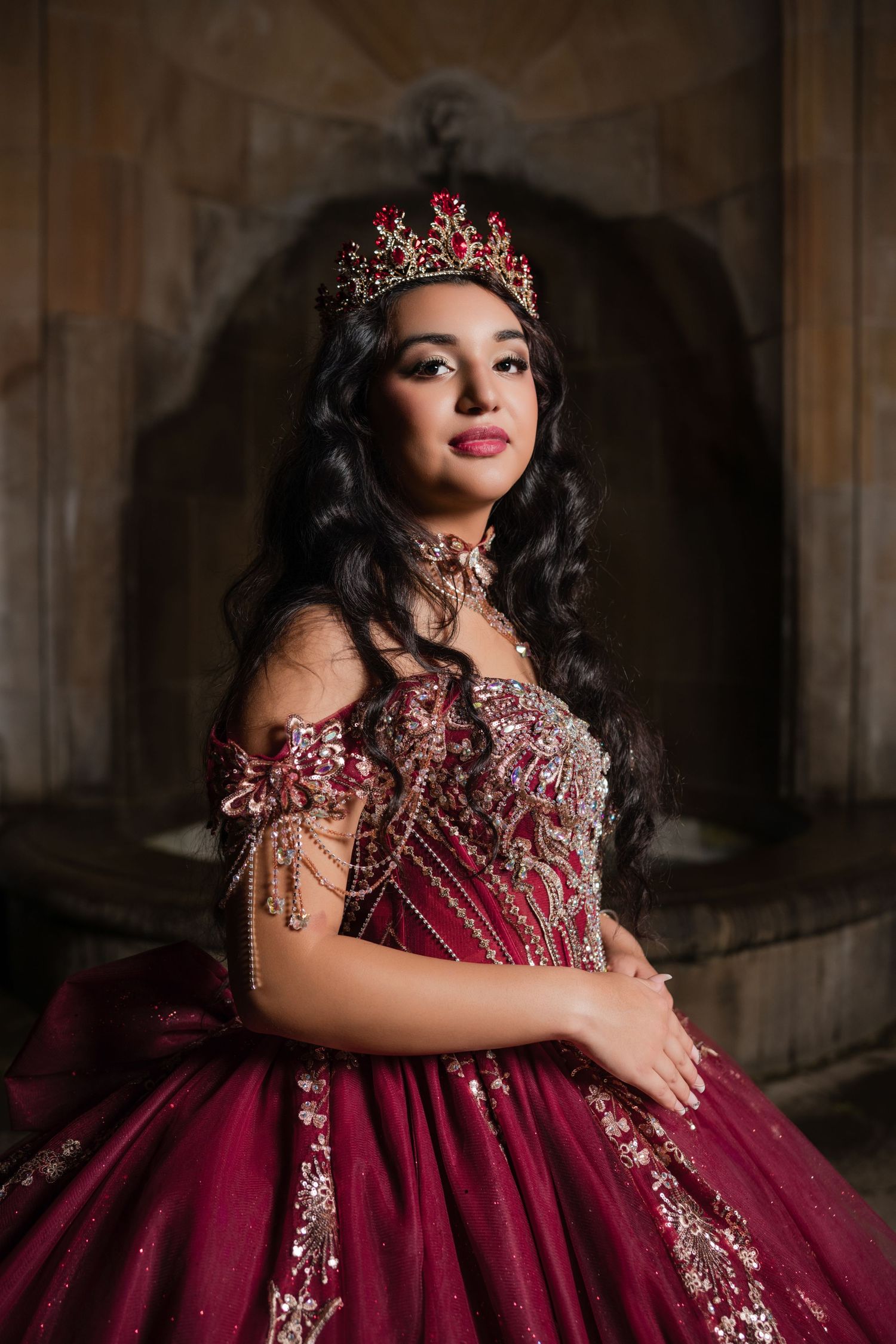 A stunning burgundy quinceanera ball gown with crystal embellishments and matching crown in a dramatic portrait setting.