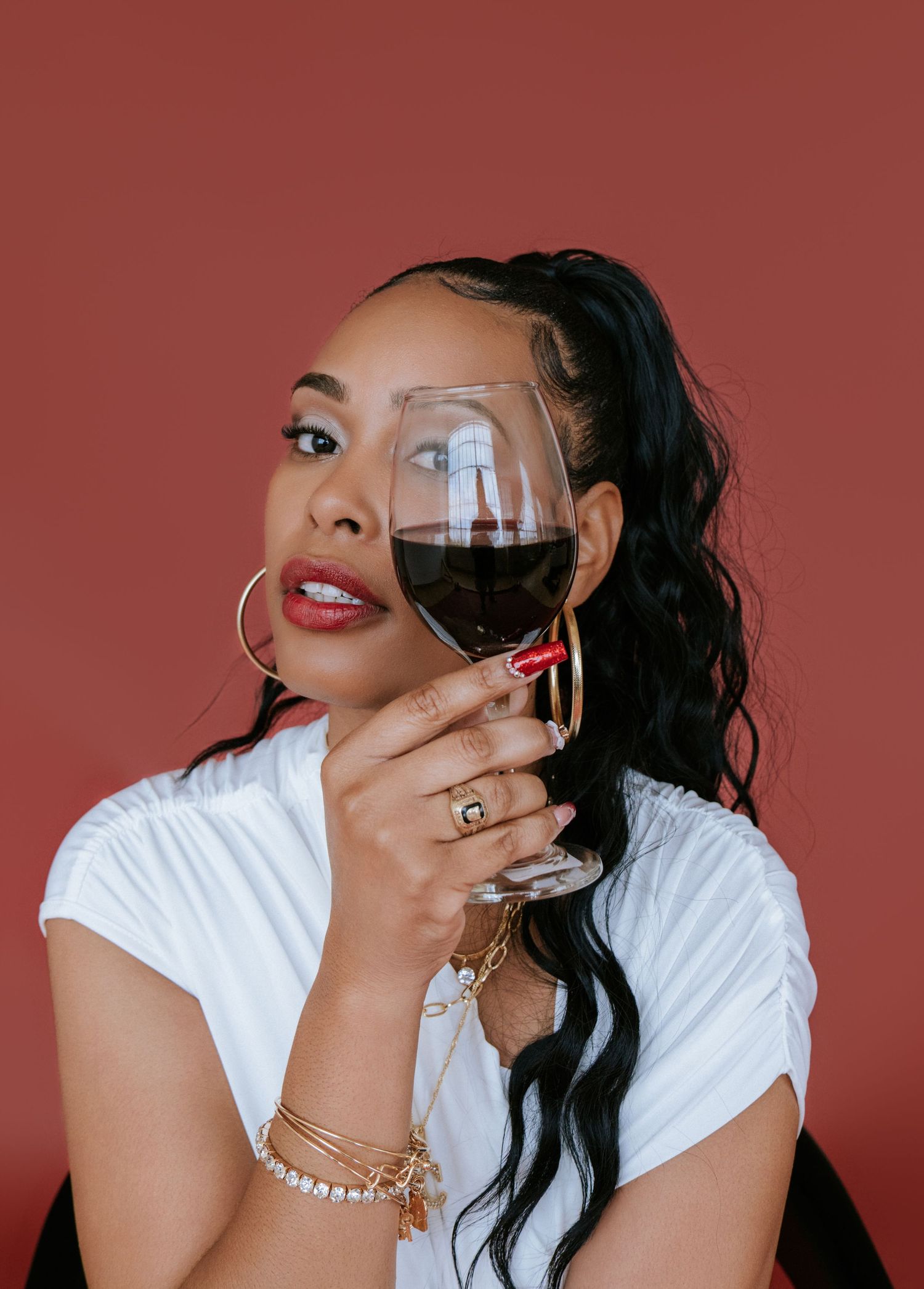 Woman in white shirt holding glass of red wine against burgundy background with hoop earrings and wavy dark hair.