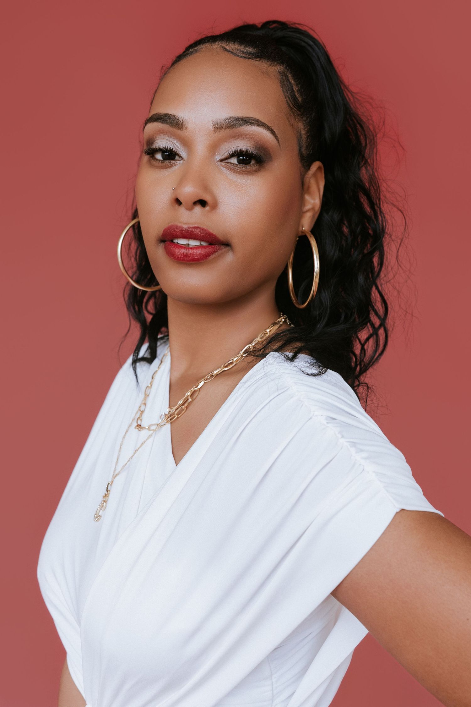 Portrait series against coral background showing glamorous styling with white top, gold hoop earrings and bold red lipstick.