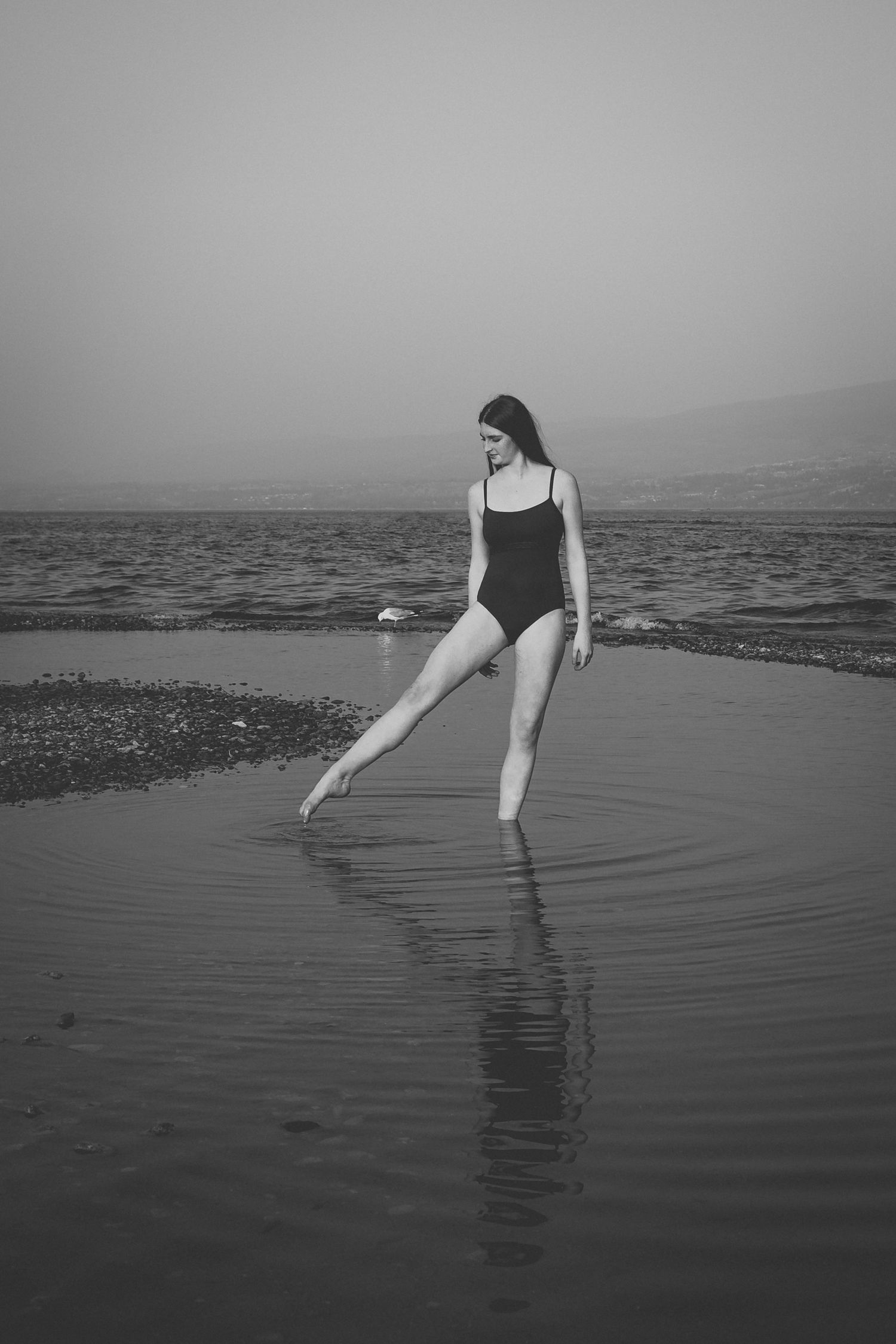 A dancer in a black swimsuit strikes an elegant pose on a misty beach, creating a reflection in the wet sand.