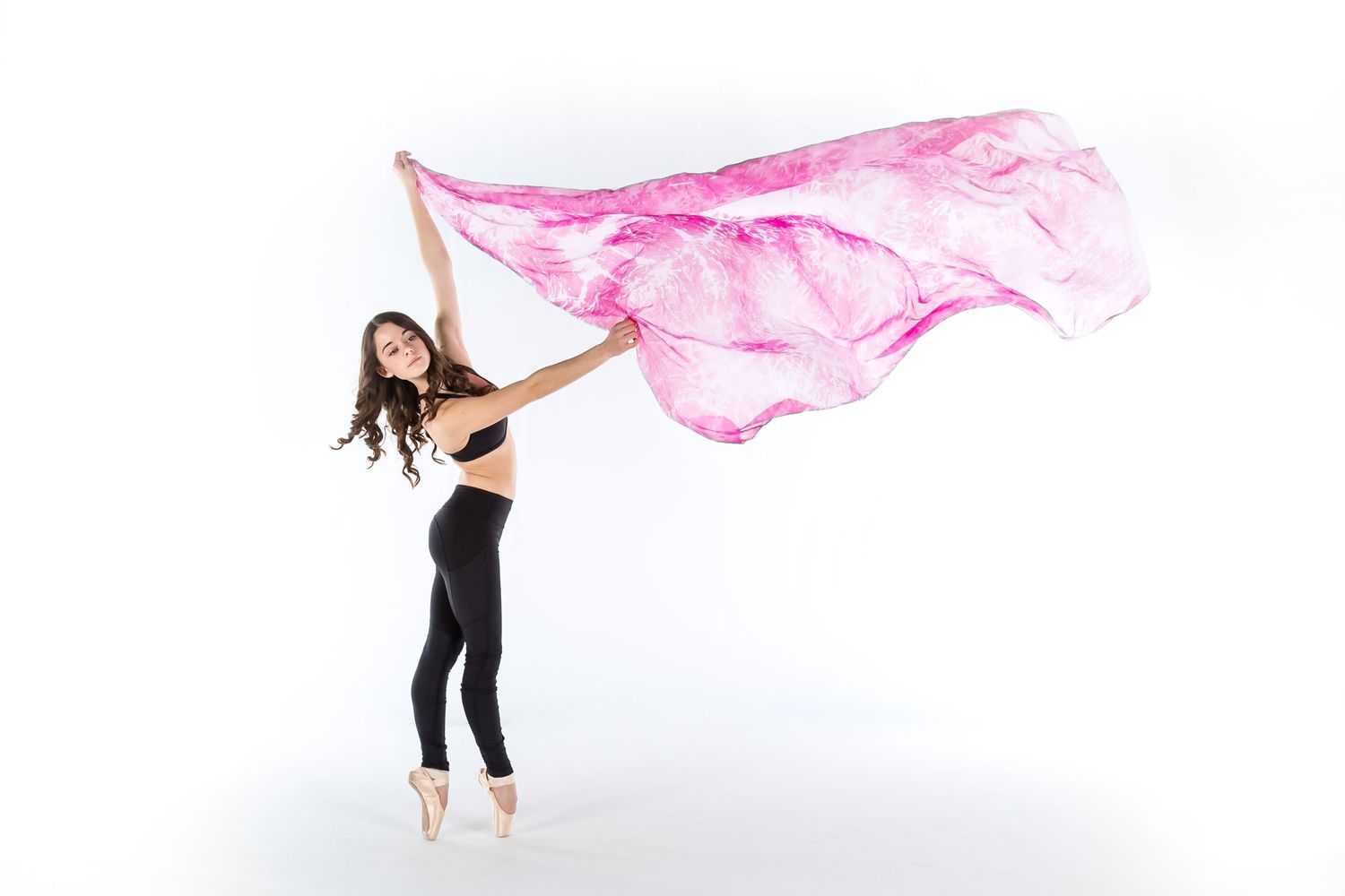 Ballet dancer in black activewear gracefully waves flowing pink silk fabric while posing en pointe against white backdrop.