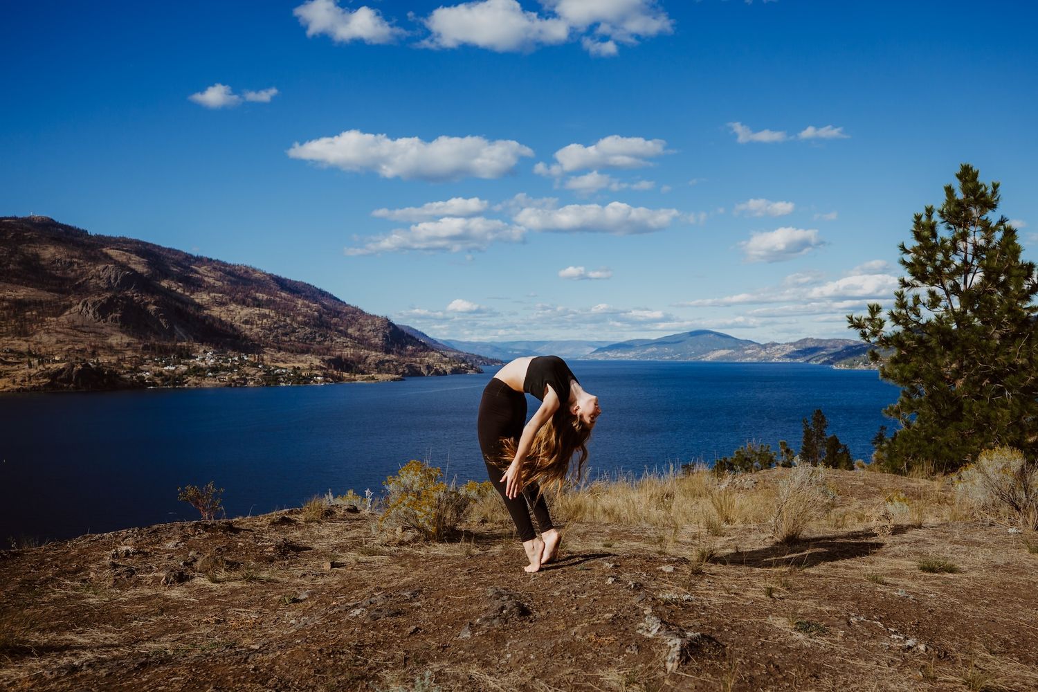 Photographer with camera equipment stands on hillside overlooking blue mountain lake on sunny day.