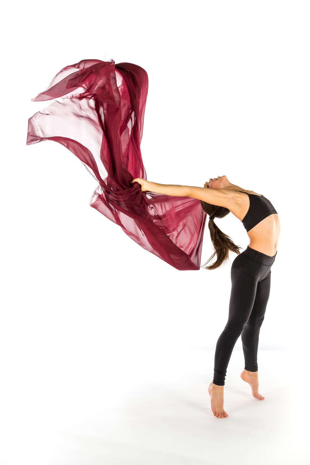 Dancer in black athletic wear bends backwards while gracefully waving a flowing burgundy fabric in an artistic pose.