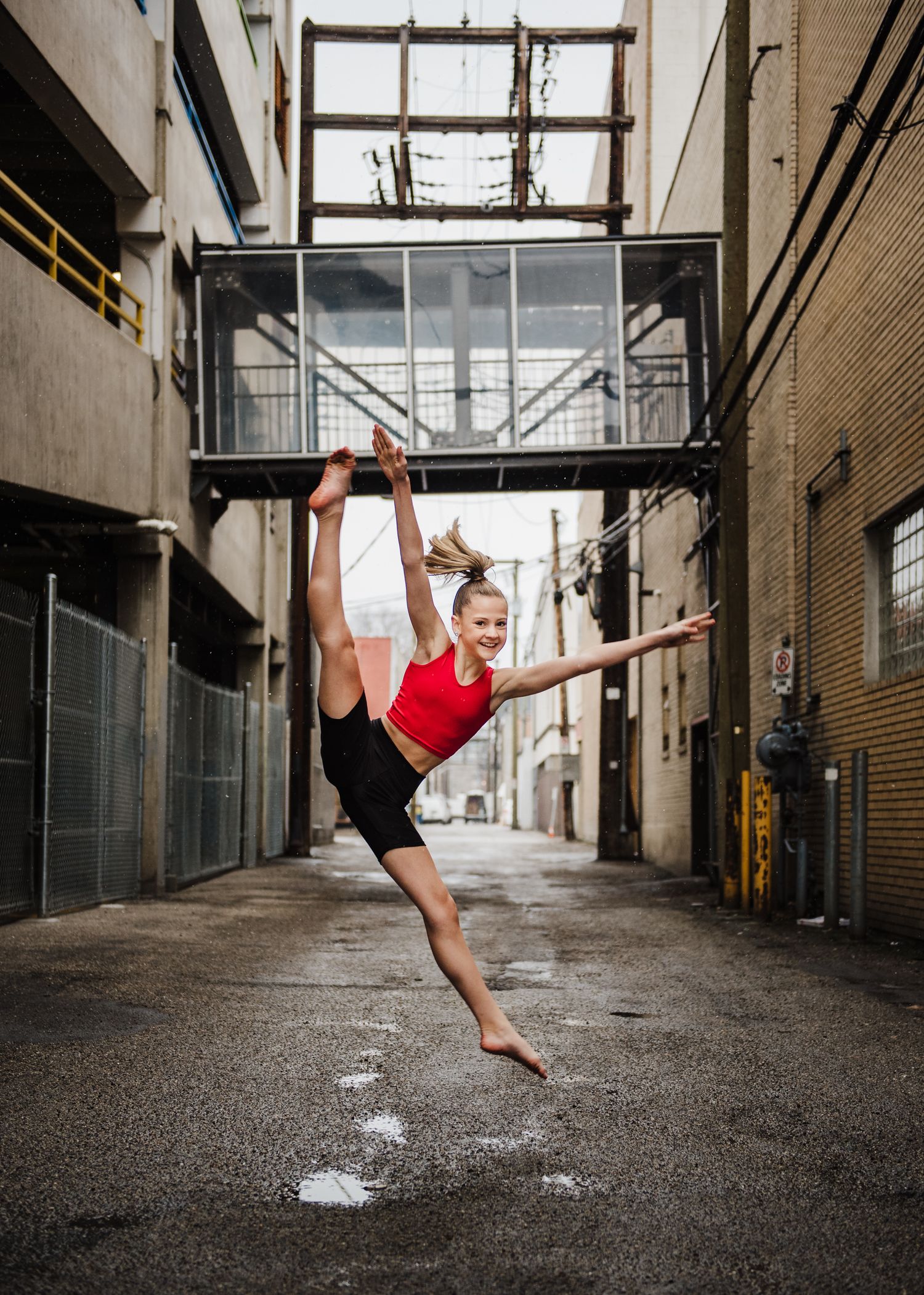 A dancer in a red top performs a dramatic split leap in an urban alley between buildings.