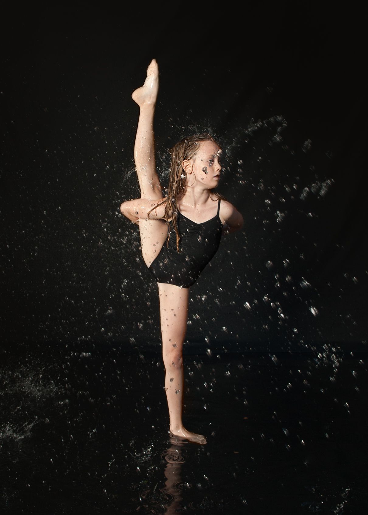 Dancer in black leotard performs dramatic leg extension pose with water splashing against dark background.