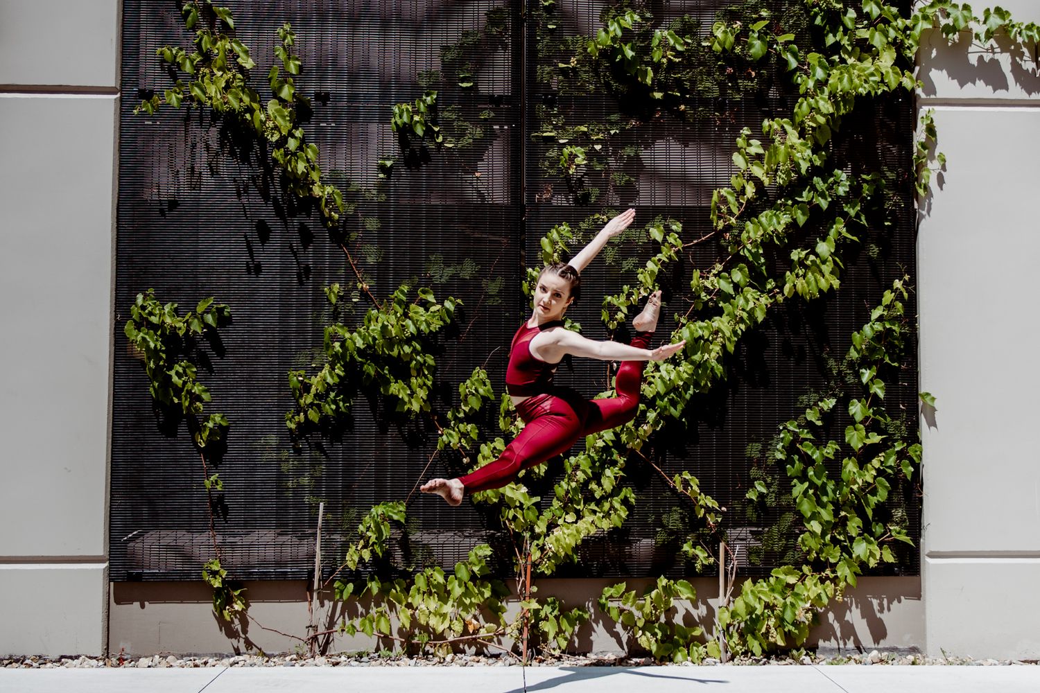A dancer in red leaps gracefully against a wall covered in climbing vines and greenery.