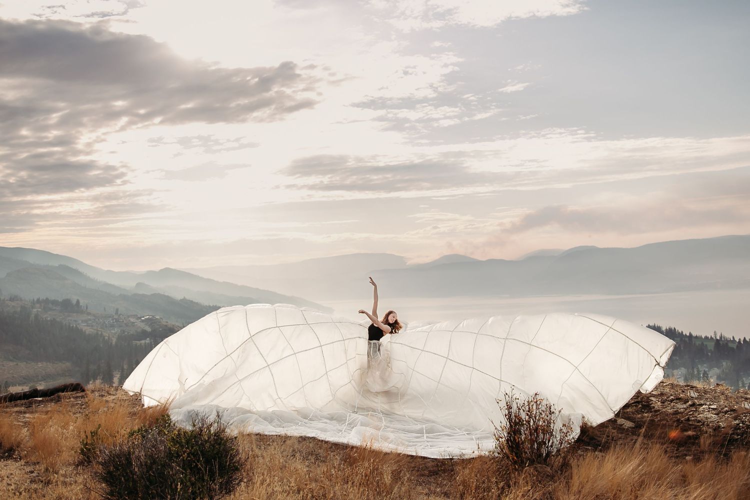 A white flowy fabric billows dramatically against a mountain landscape at sunset with a person emerging from beneath.