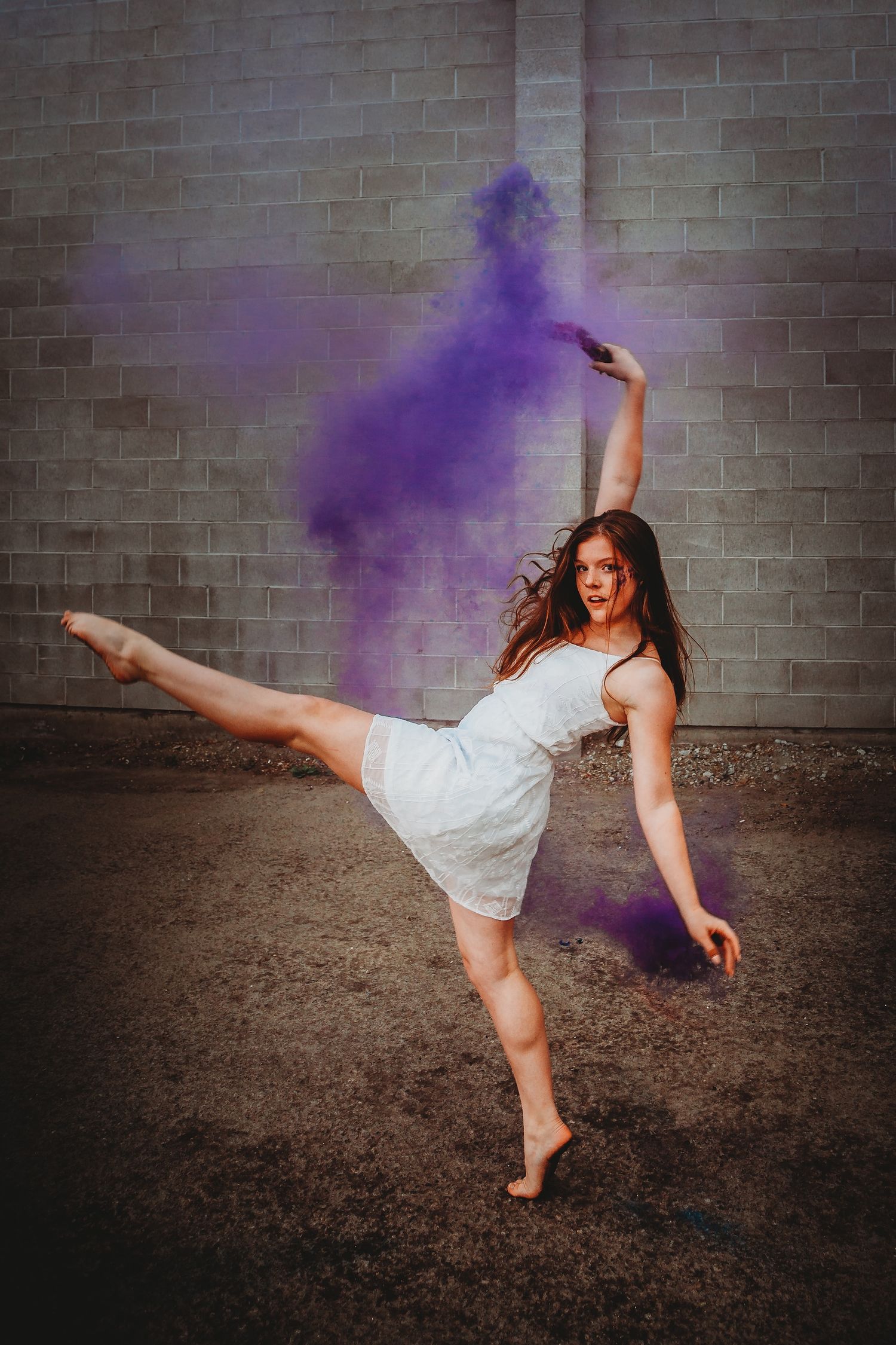 A dancer in a white dress performs a dynamic pose against a brick wall with purple powder creating artistic smoke effects.