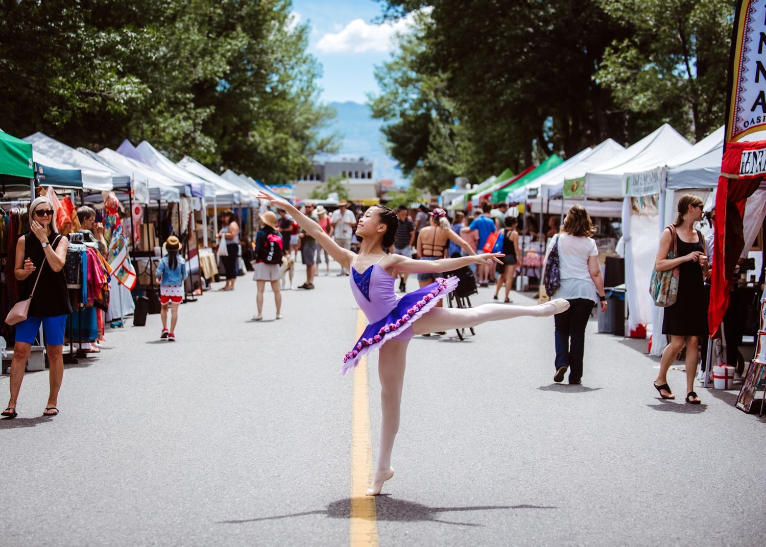 People browse vendor booths at an outdoor street festival while a person in the center does a ballet-like pose.