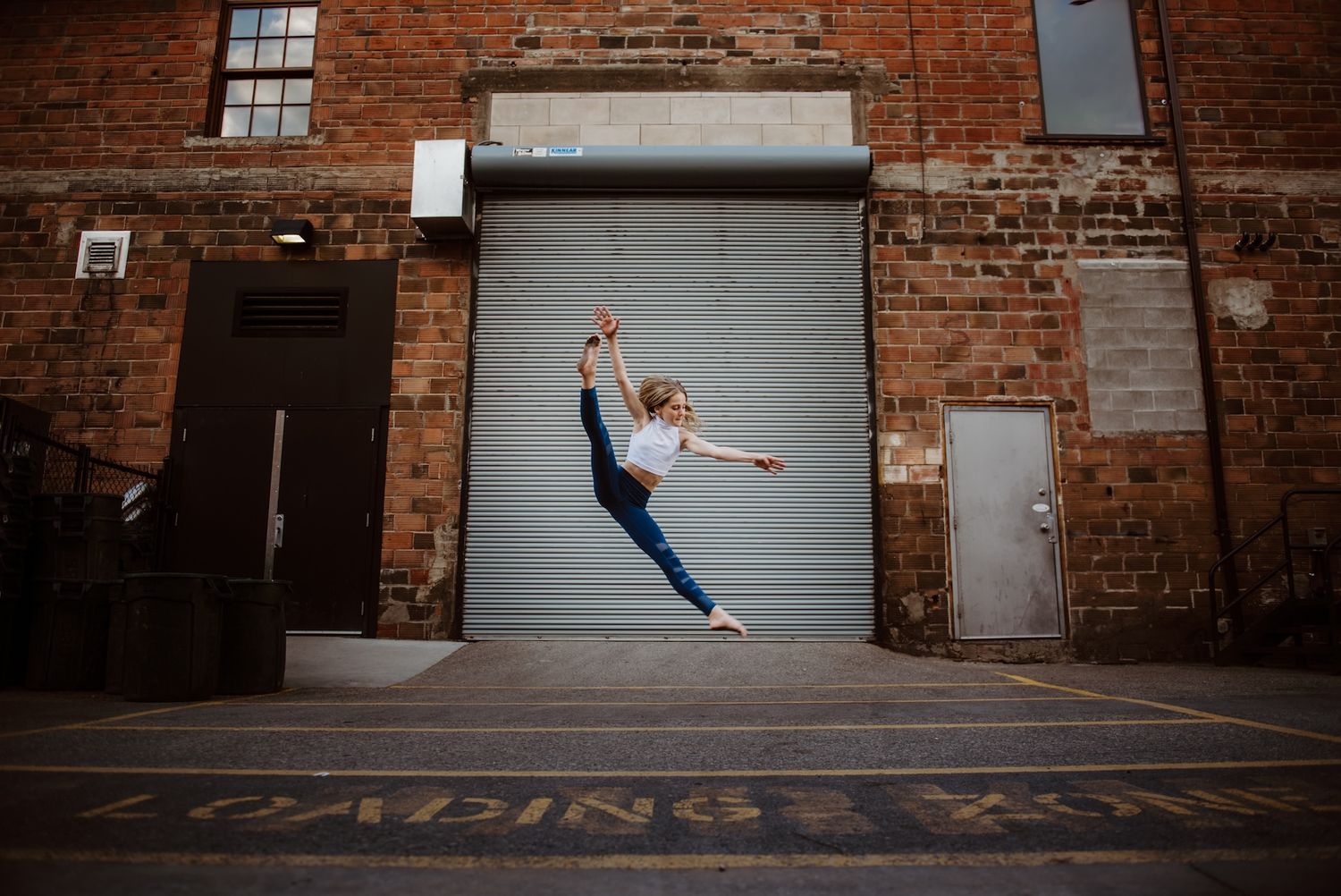 A dancer leaps gracefully in front of a rolling metal door on a brick industrial building.