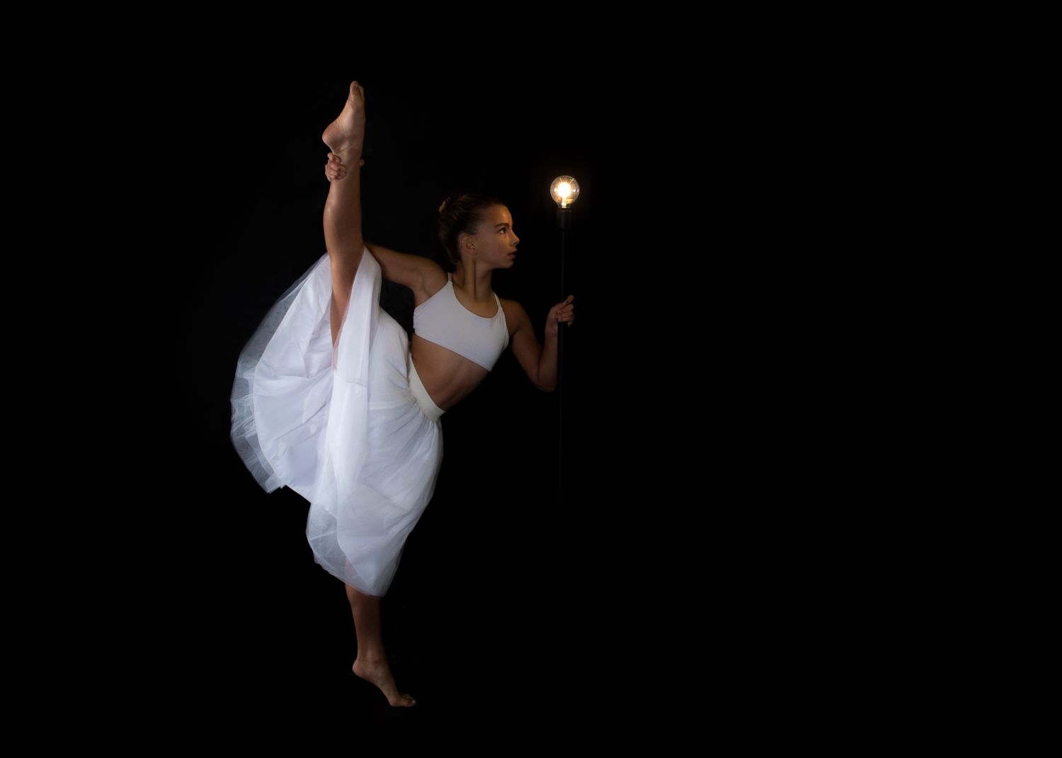 A dancer in white performs a dramatic high kick against a dark background with a glowing light source.