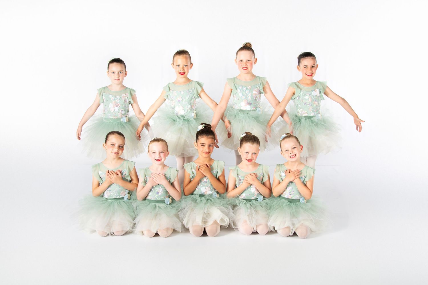 A dance group in matching mint green tutus poses in two rows on white backdrop for a ballet performance photo.