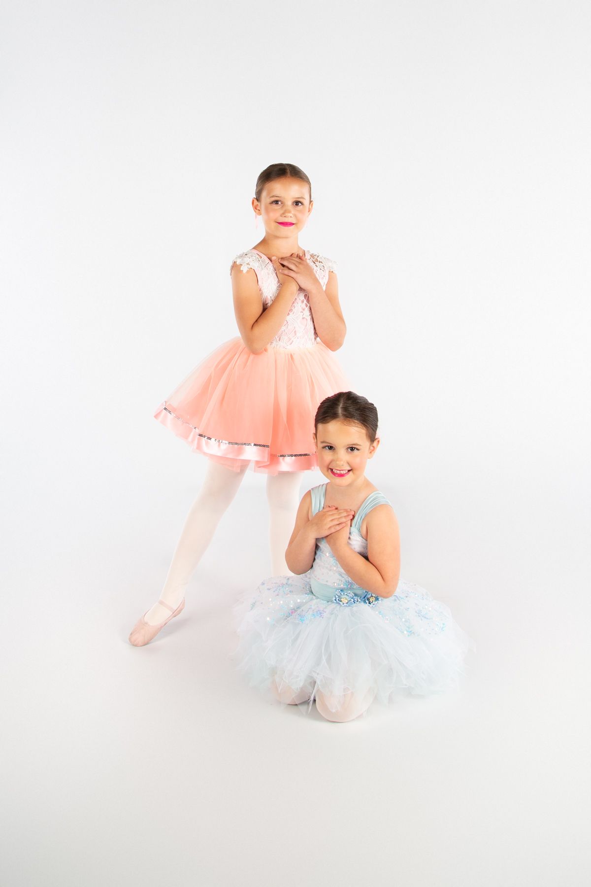 Two dancers in pastel tutus pose gracefully in studio against white background.