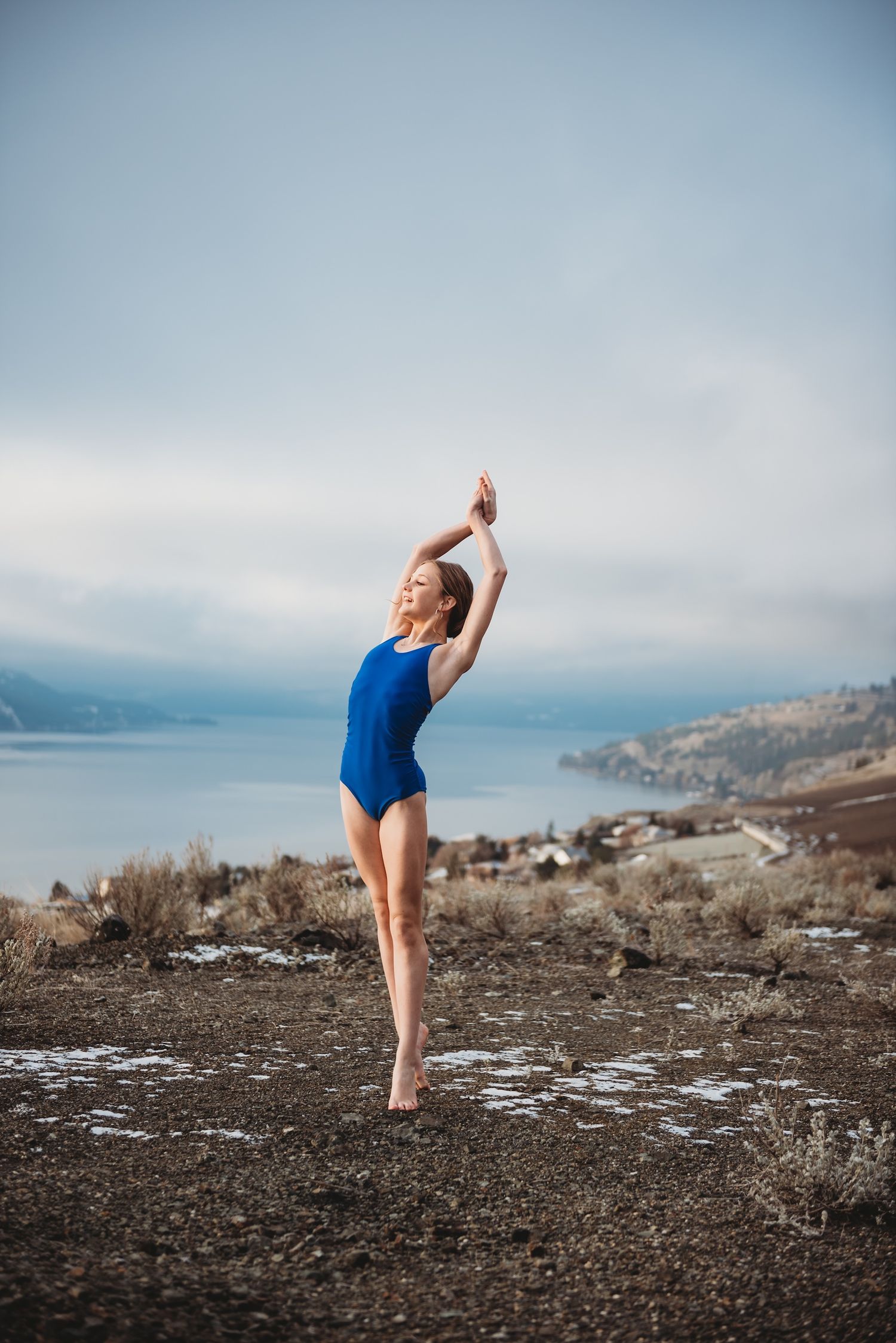 Dancer in blue leotard performs graceful pose on mountainside overlooking lake and cloudy landscape.